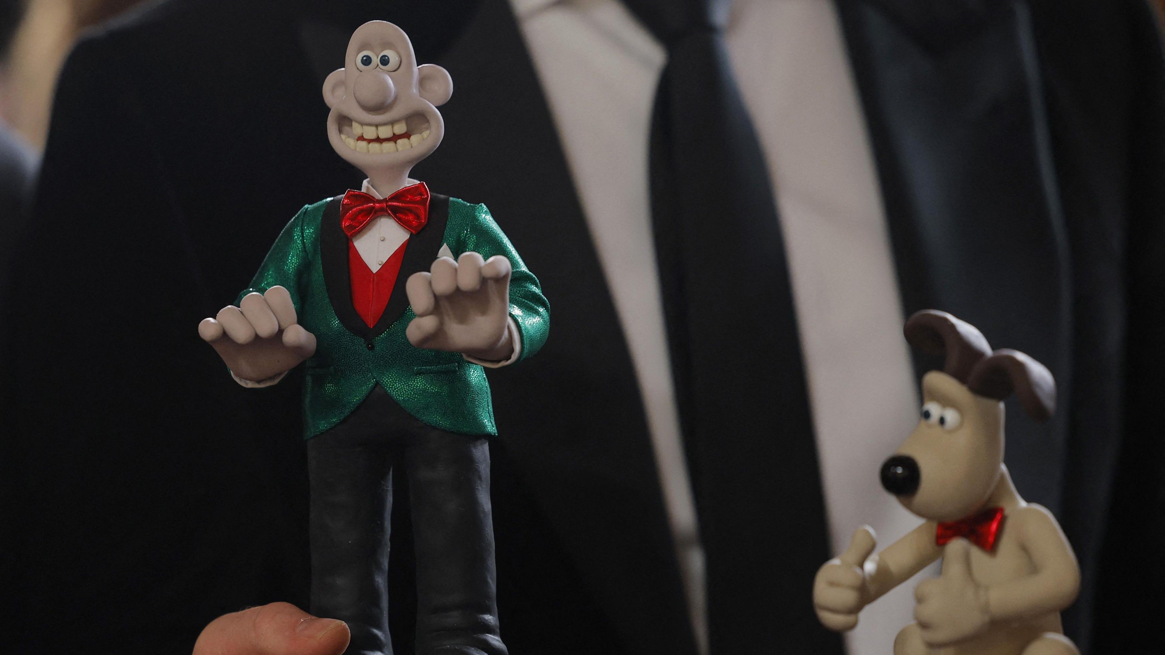 A man wearing a dark suit and tie holds two small models of Aardman characters Wallace & Gromit