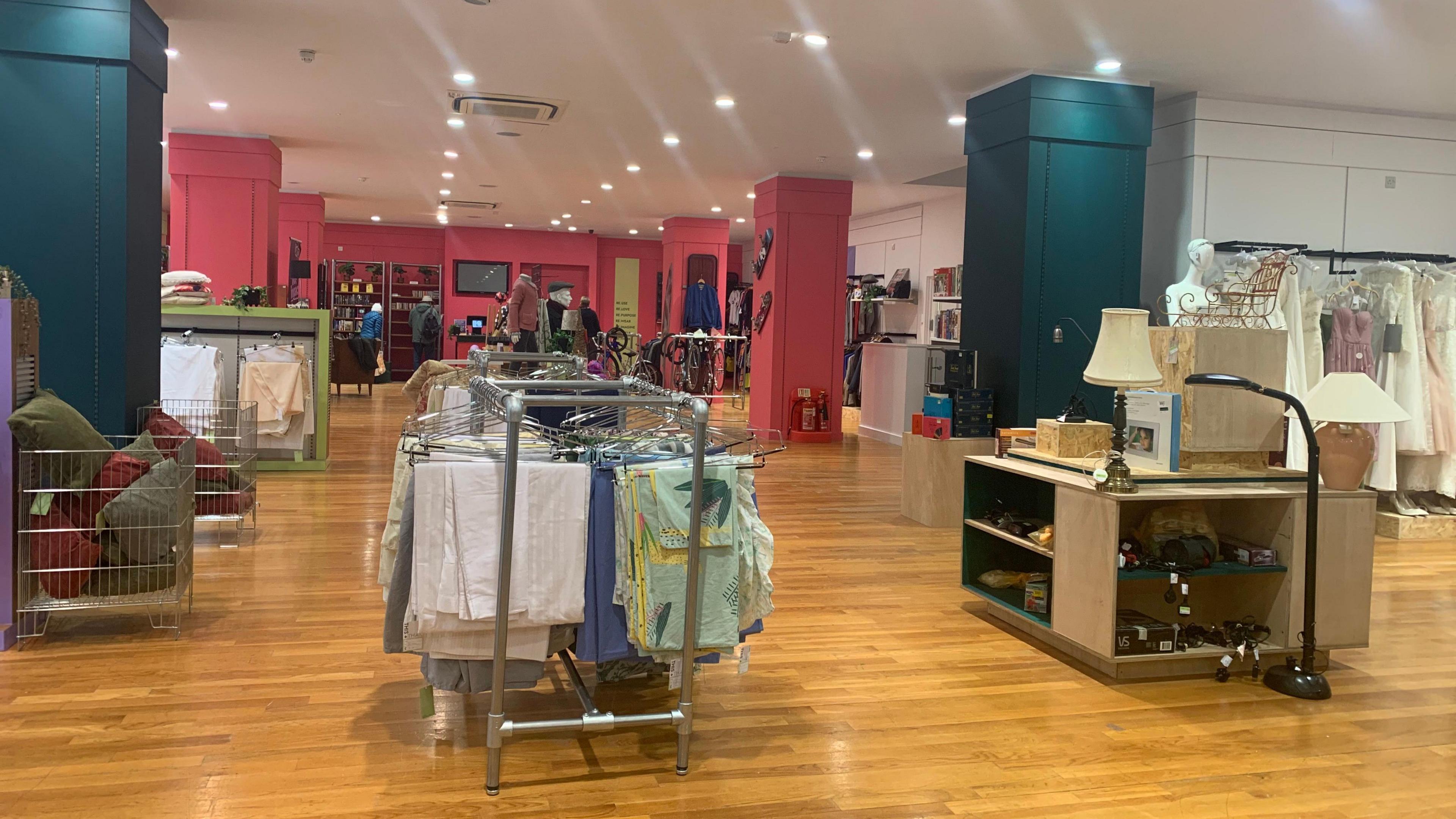 A shop interior with a wooden floor, teal and pink supporting pillars and racks of clothes.