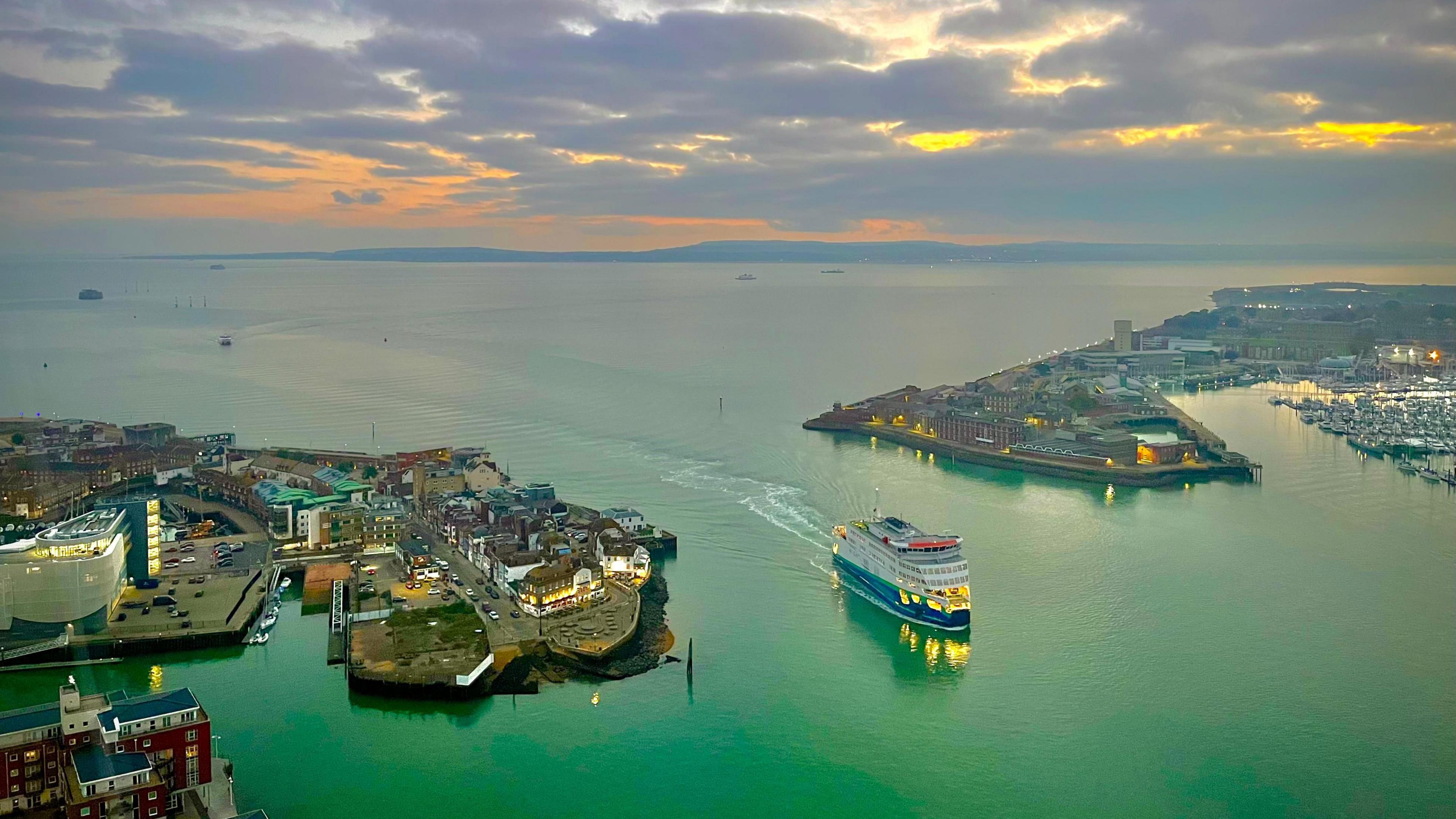 The sunset over the Solent with the Isle of Wight in the distance and an orange sky glowing through the clouds. In the foreground, a ferry can be seen coming through the entrance to Portsmouth Harbour. It is twilight and the buildings on each side of the harbour have lights on and the water is a deep green colour. 