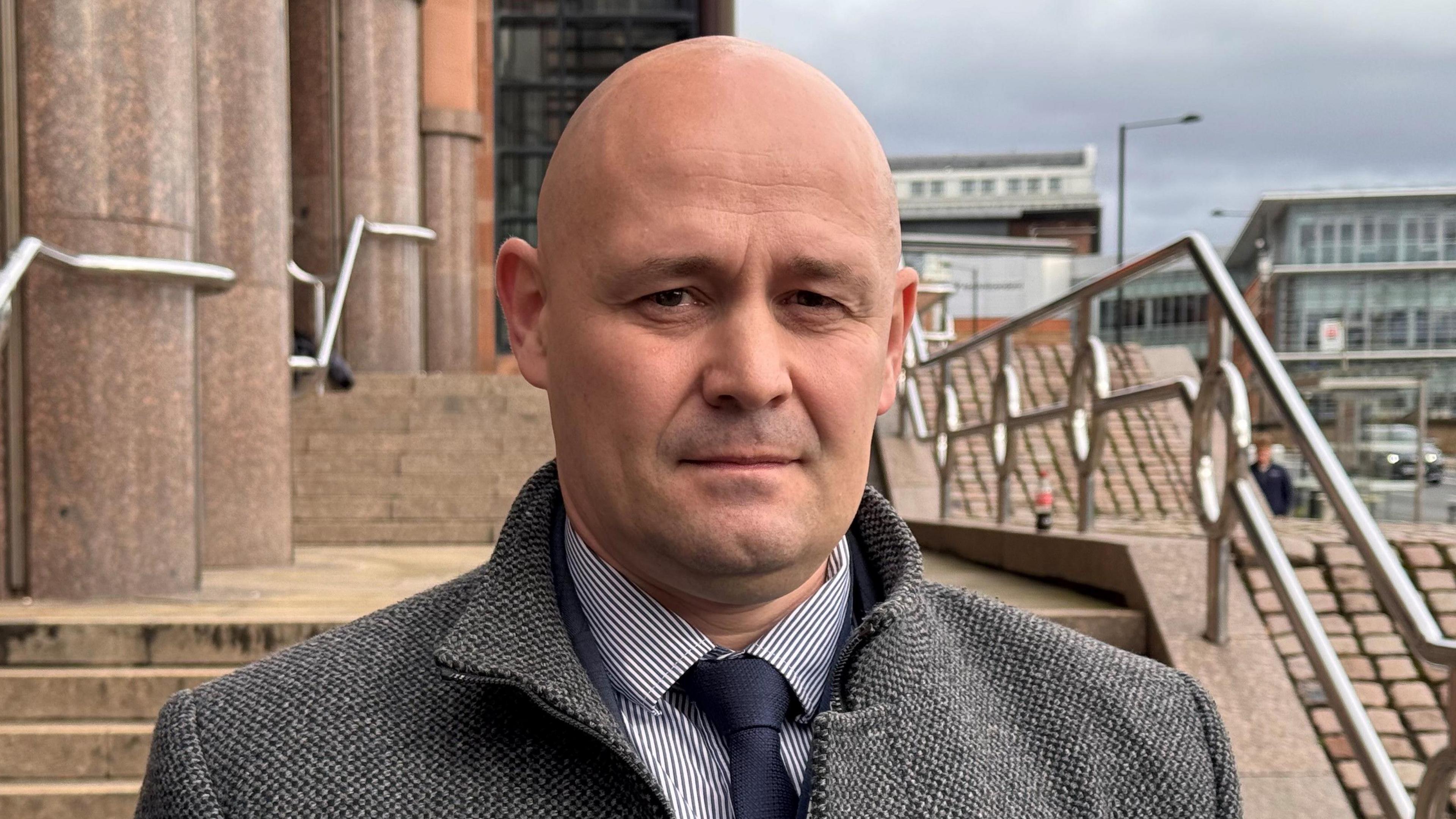 Det Ch Insp Jason Henry stands outside the court building, He is bald and is wearing a grey coat, white and navy striped shirt and a navy tie.