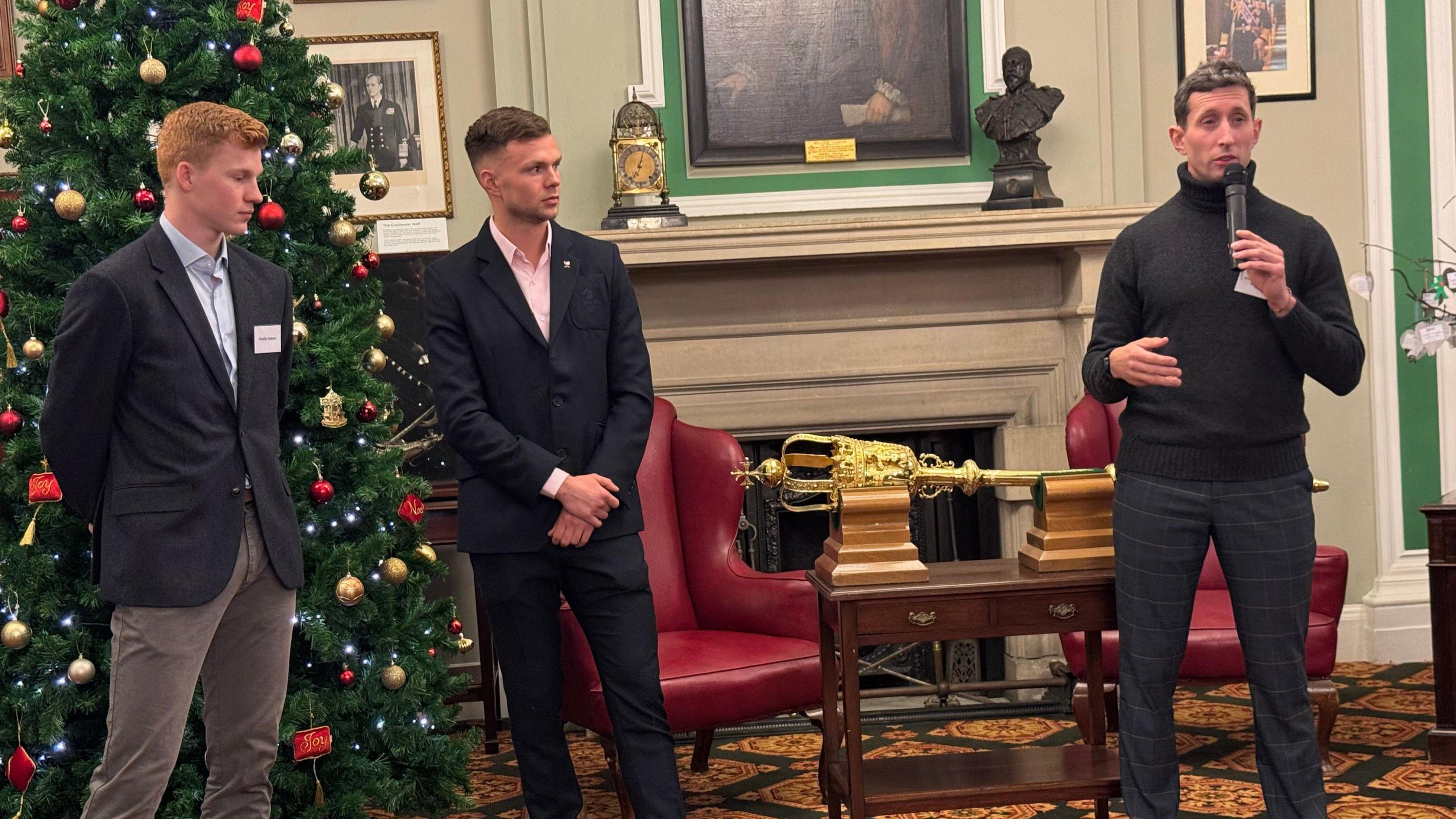 Three men standing in front of a Christmas tree and two red leather chairs. Two of the men are wearing suits and the third man is holding a microphone and wearing a black turtleneck jumper.