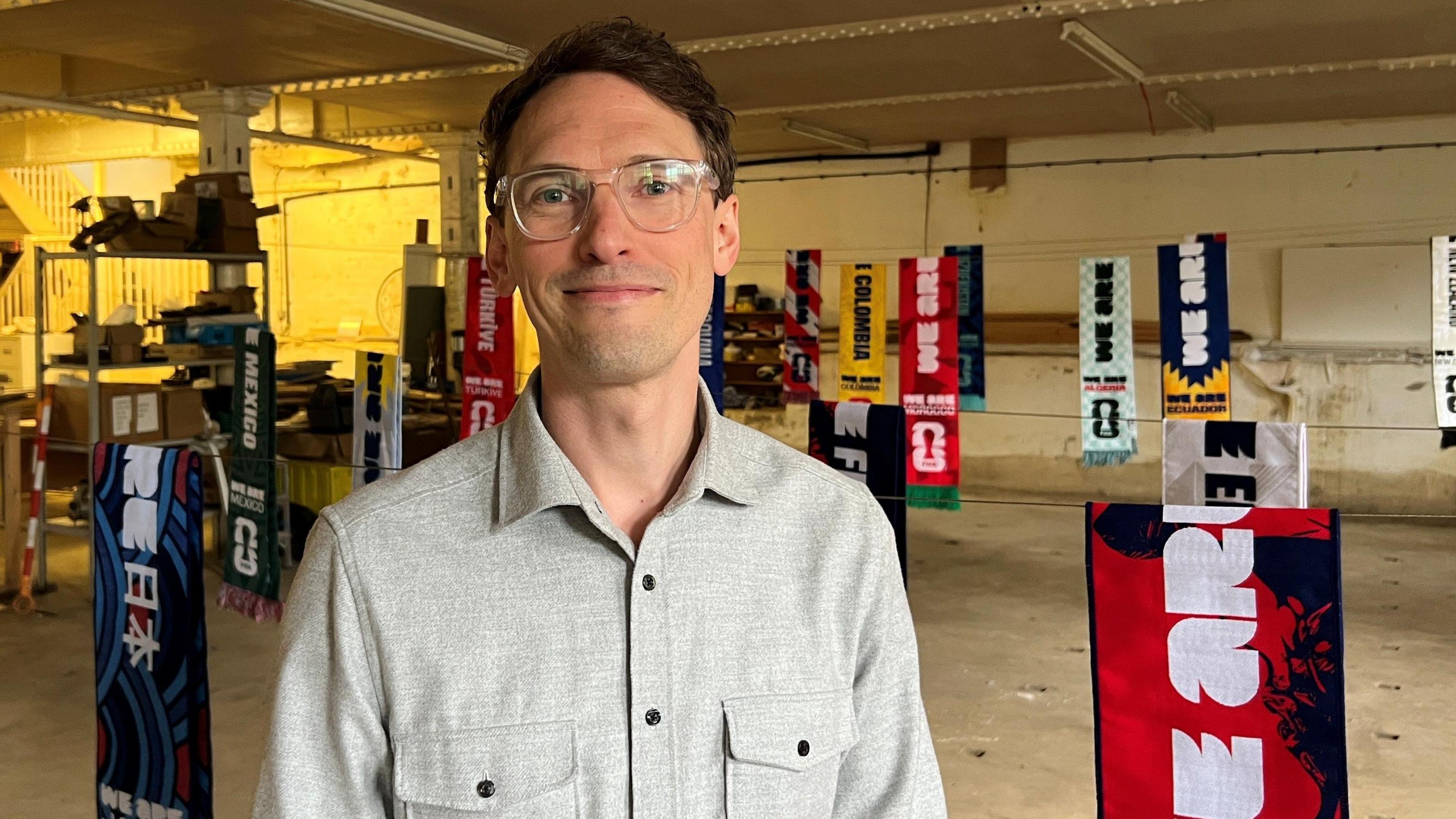 A picture of Tom Pitts, who has dark brown hair and glasses, standing in front of a display of his World Cup scarves.