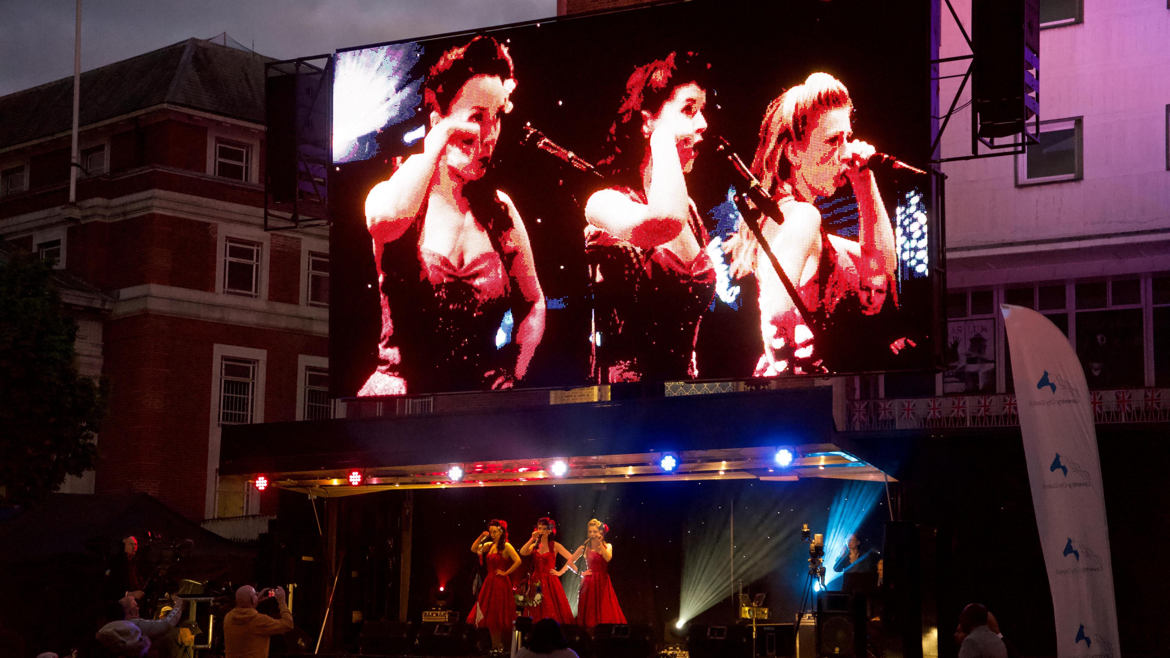 A giant screen above the stage showing three singers in red 1940s dresses