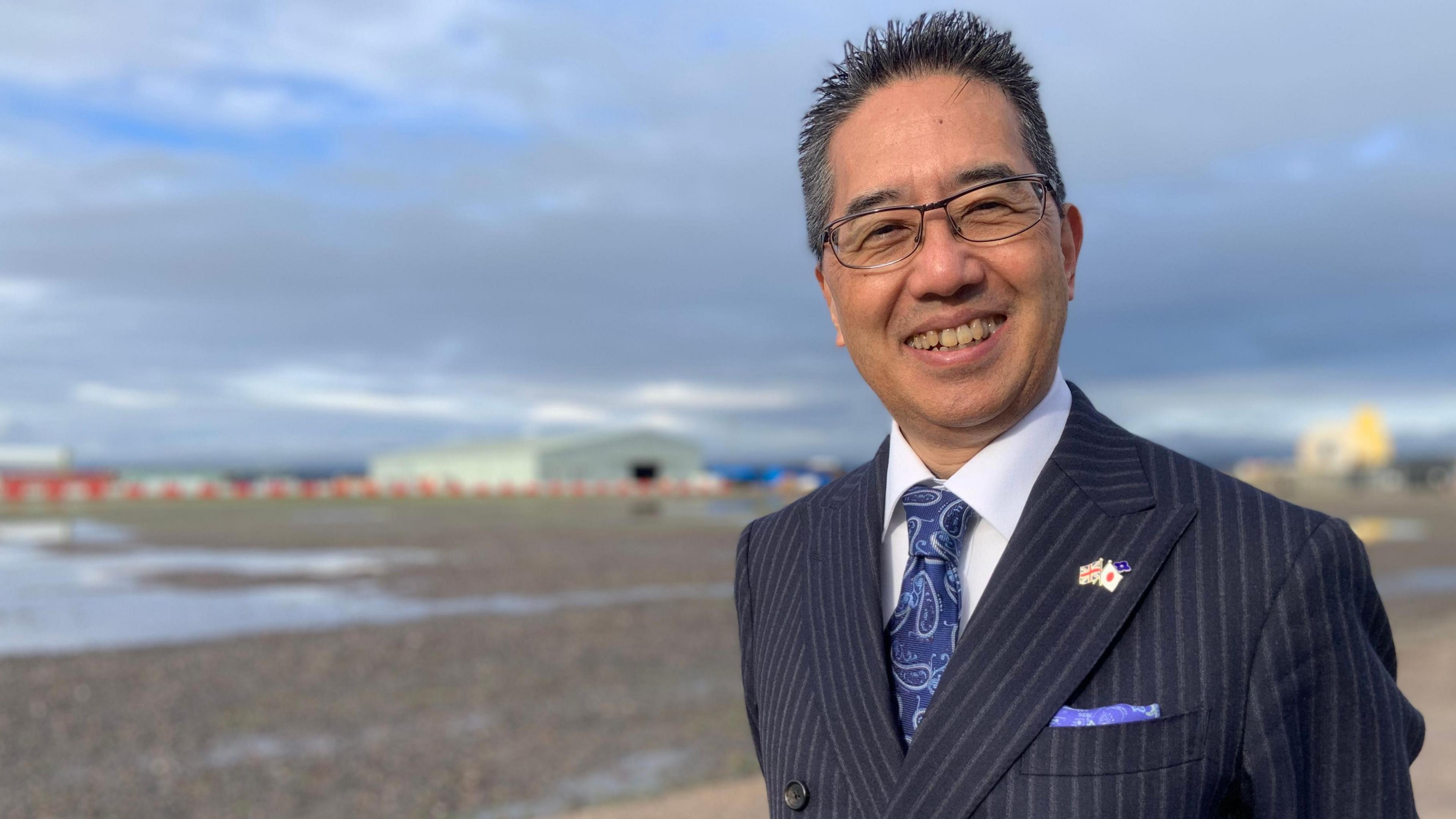 Yasyuki Shibata smiles at the camera. He is wearing a navy pin stripe suit, white shirt and Paisley-patterned tie. He has dark spiky hair and black-rimmed. glasses.
