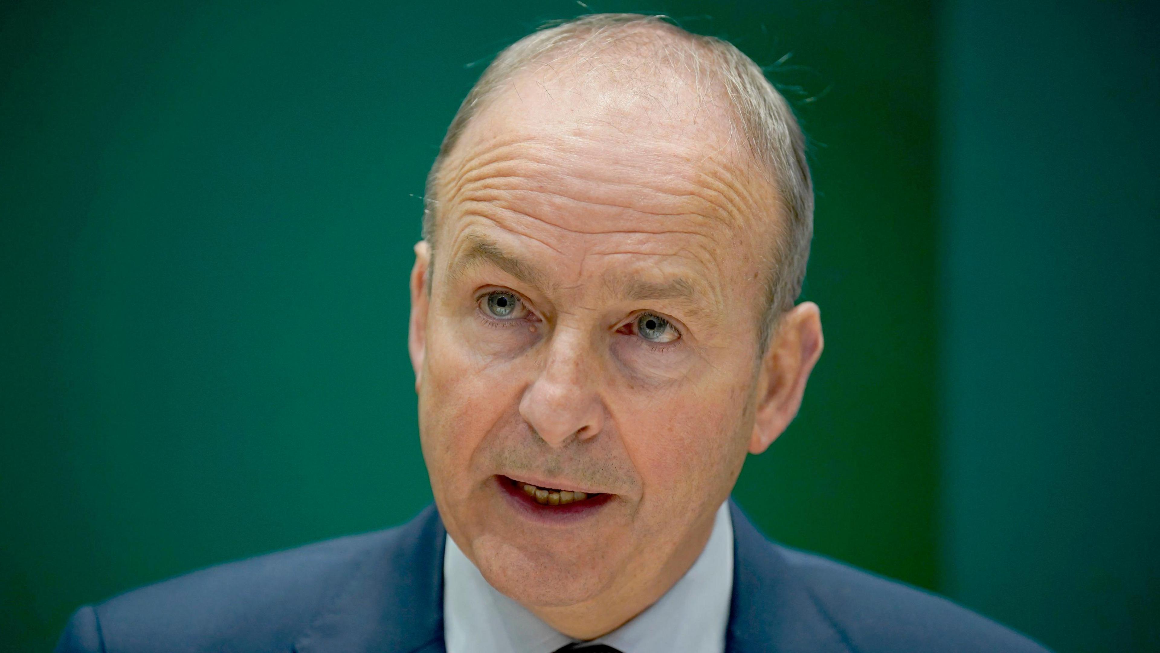 Micheál Martin - a man with grey hair is speaking and looking to the left of the camera. He is in front of a blurred green background and is wearing a navy suit jacket, a light collared shirt and a black and red spot tie.