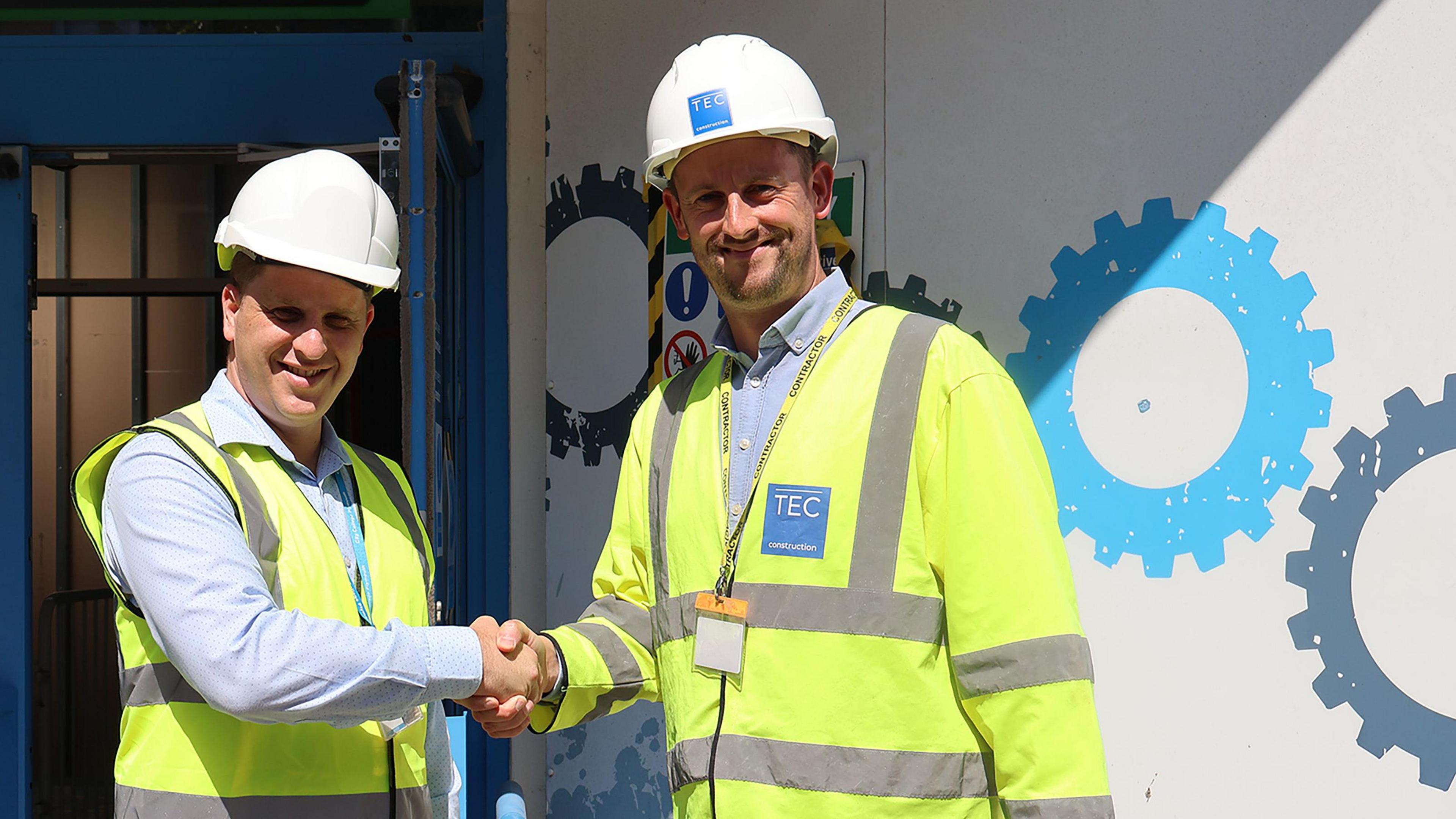 Dominic Jennings of City College Plymouth shaking hands with James Carson of TEC Construction, both of them are wearing white hard hats and yellow fluorescent jackets