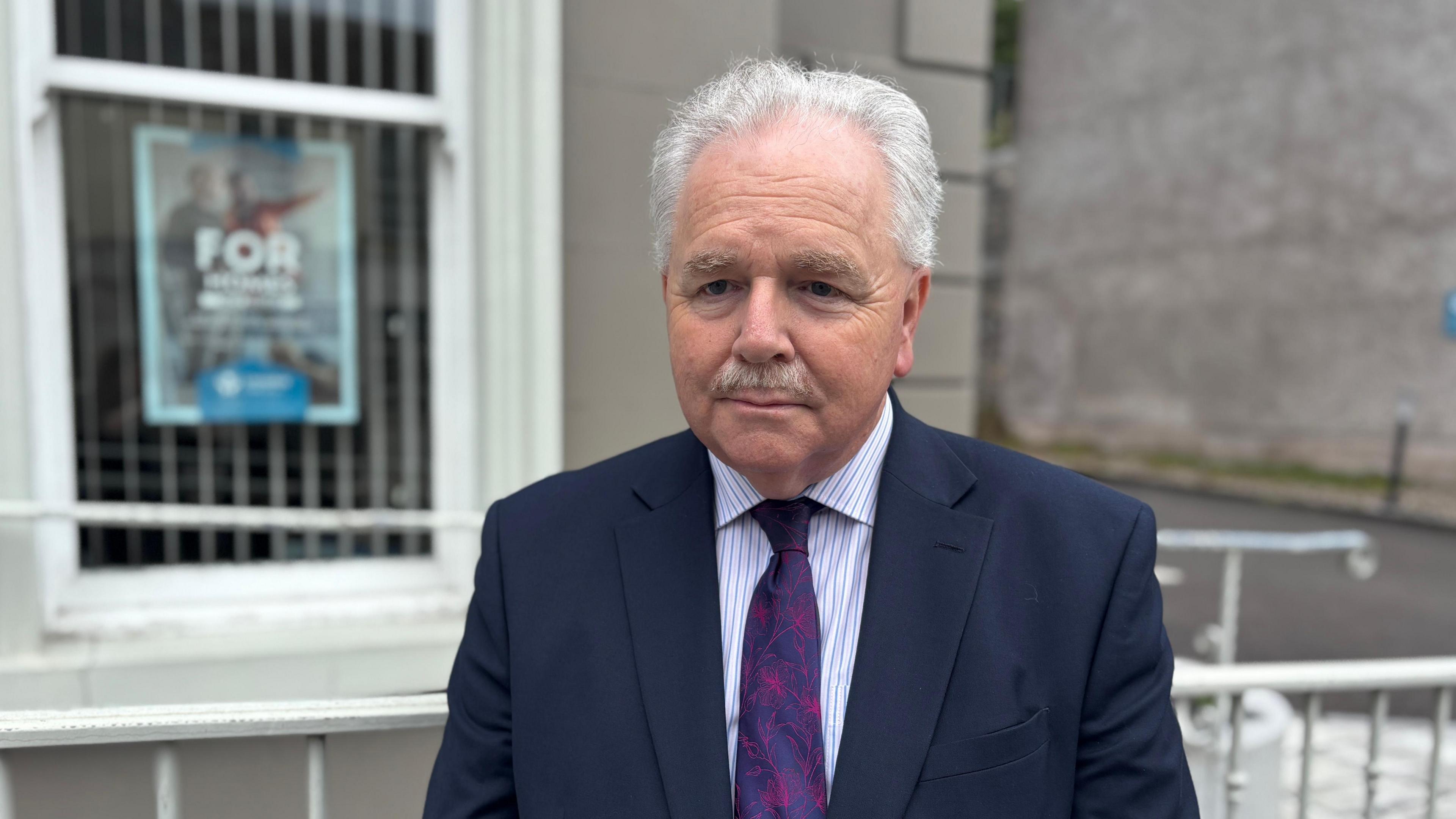A smartly dressed man stands outside a building. He has grey hair and a mustache. He is well dressed in a navy suit with a striking purple print tie.