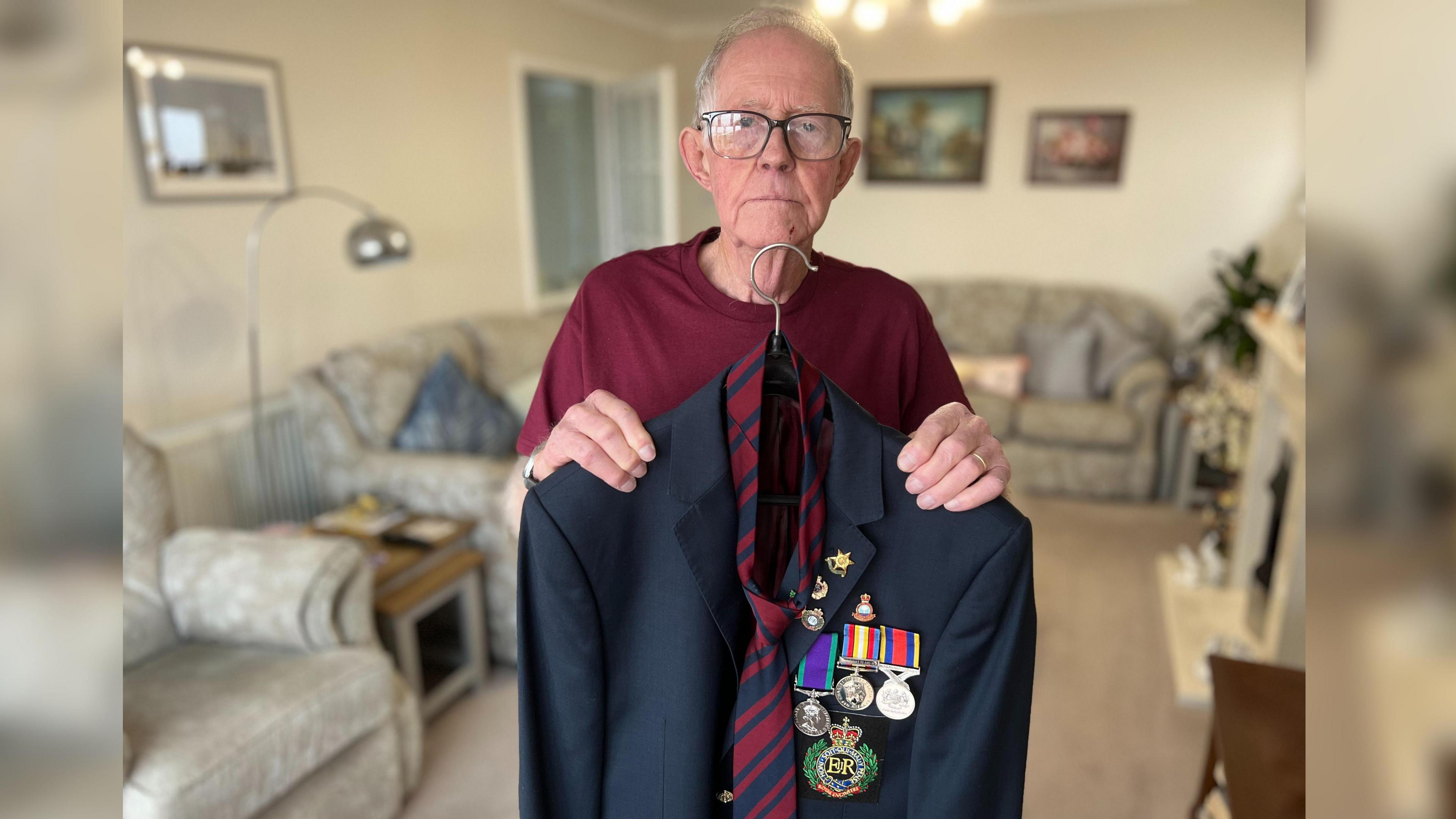 Ray Peace stands in his living room and he is holding a suit and tie on a hanger in front of his body. The suit jacket has medals on it while in the background you can see his living room which has light grey sofas and pictures on the wall.