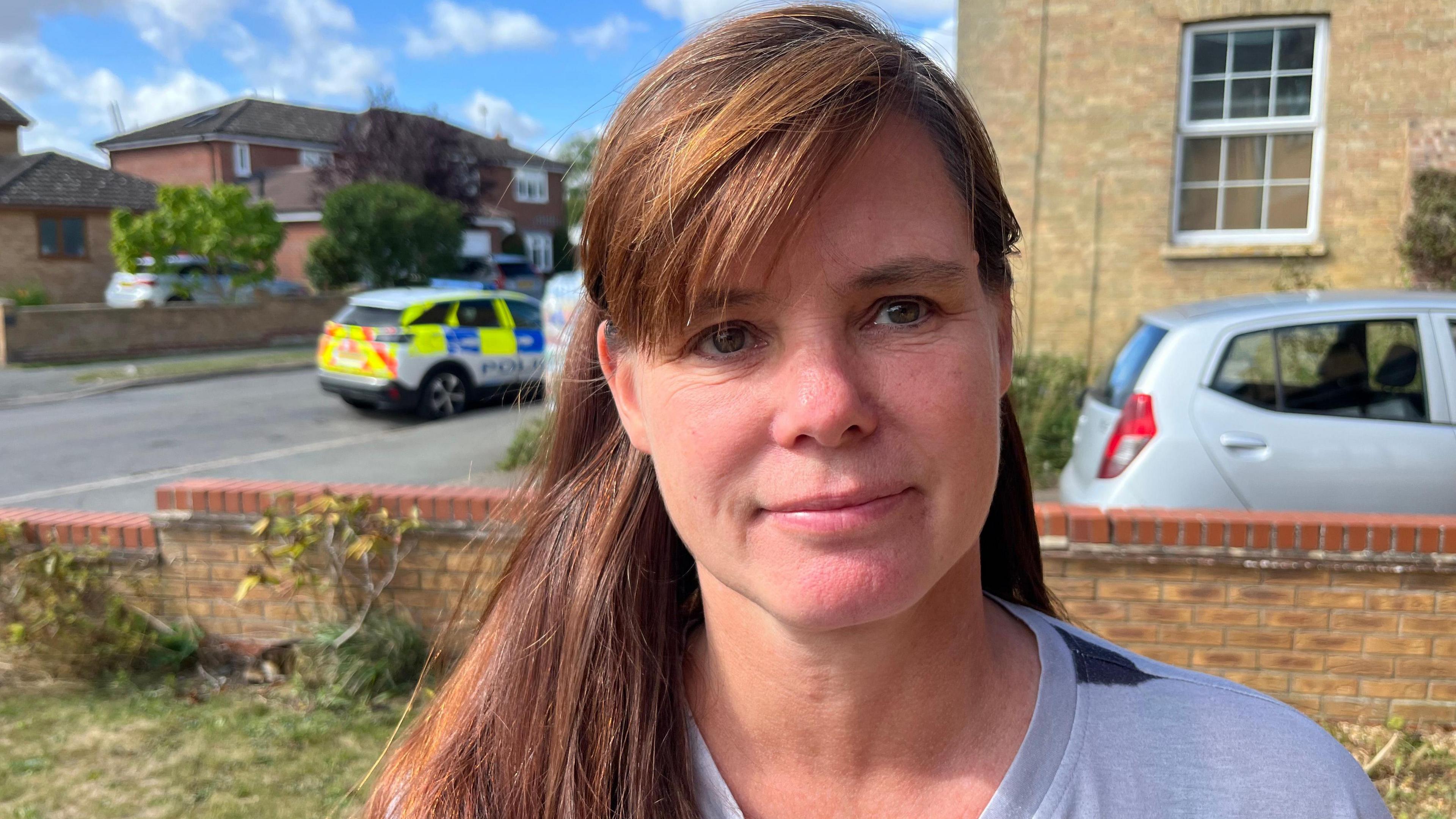 Ms Gibbs is wearing a light coloured top and is standing to the side of a house during the day. She has long brown hair and is smiling. Behind her is a parked police car.