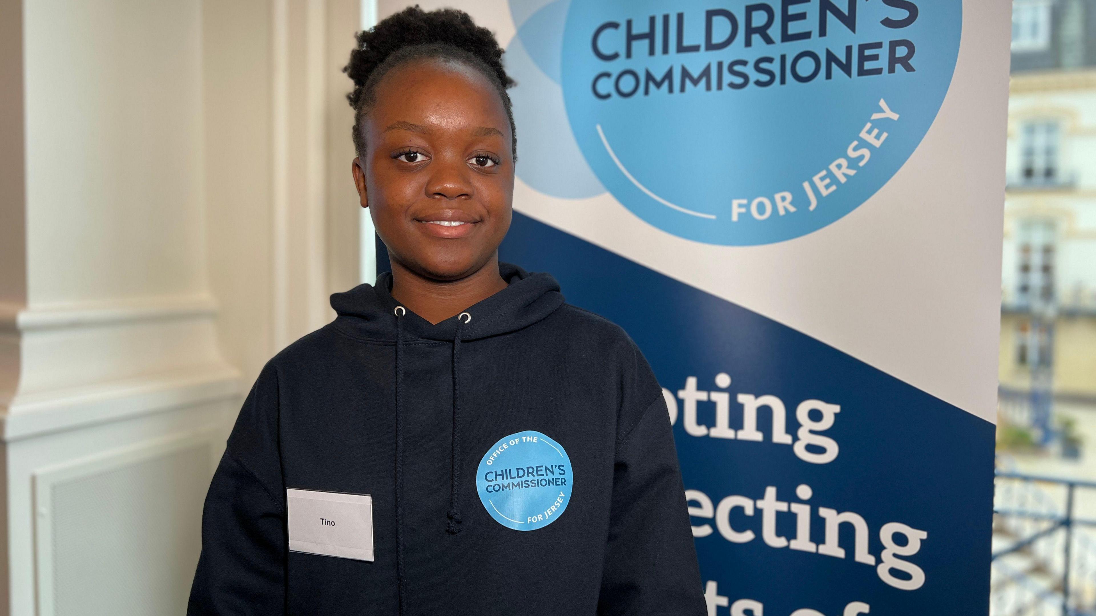 Tino smiles at the camera as she stands by a sign about the Jersey Commissioner's office. She's wearing a dark blue hoodie and her hair is tied back.