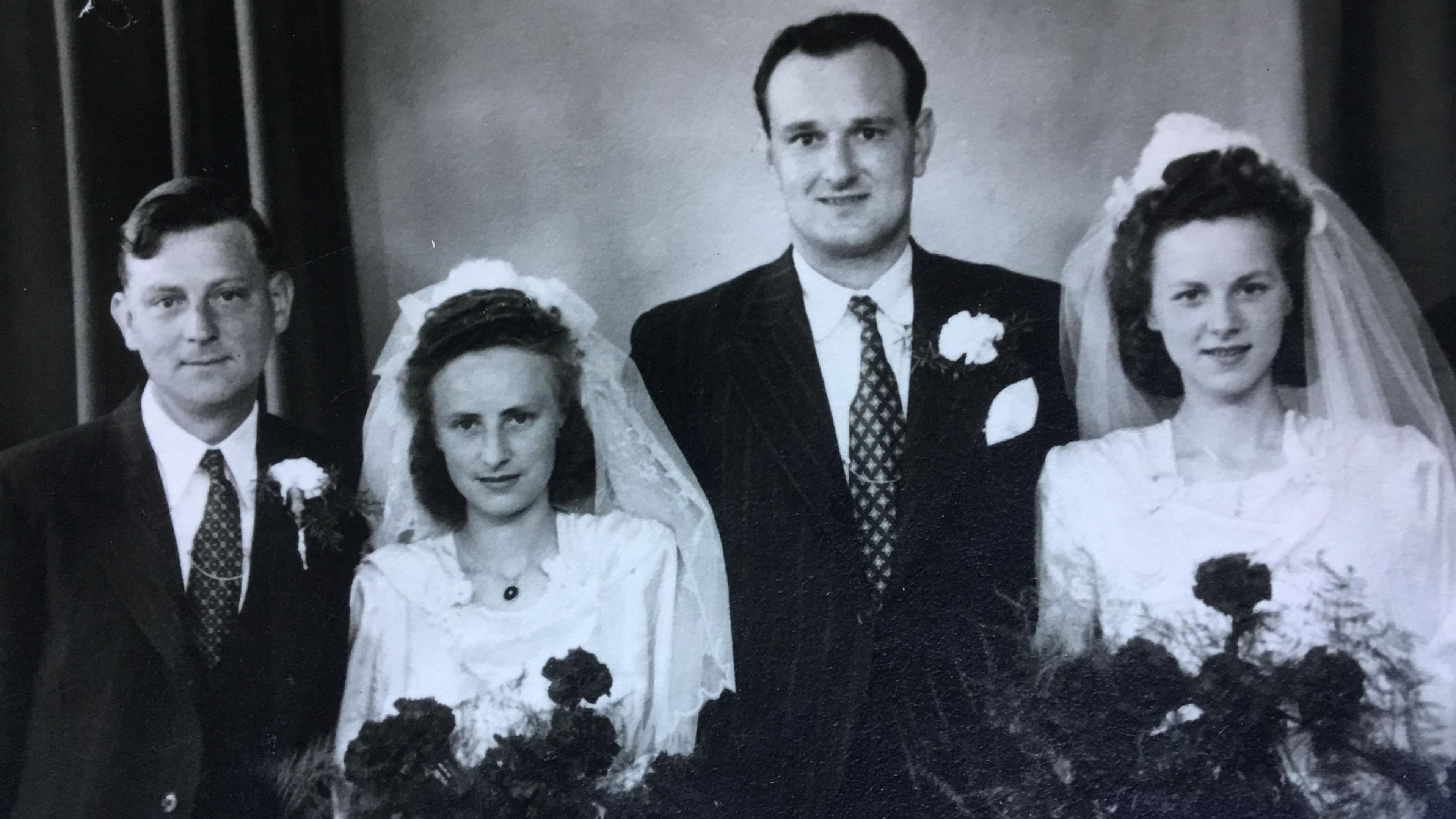 A black and white photograph of Joan and her sister Monica on their wedding day, dressed in bridal gowns and veils, standing alongside their new husbands.