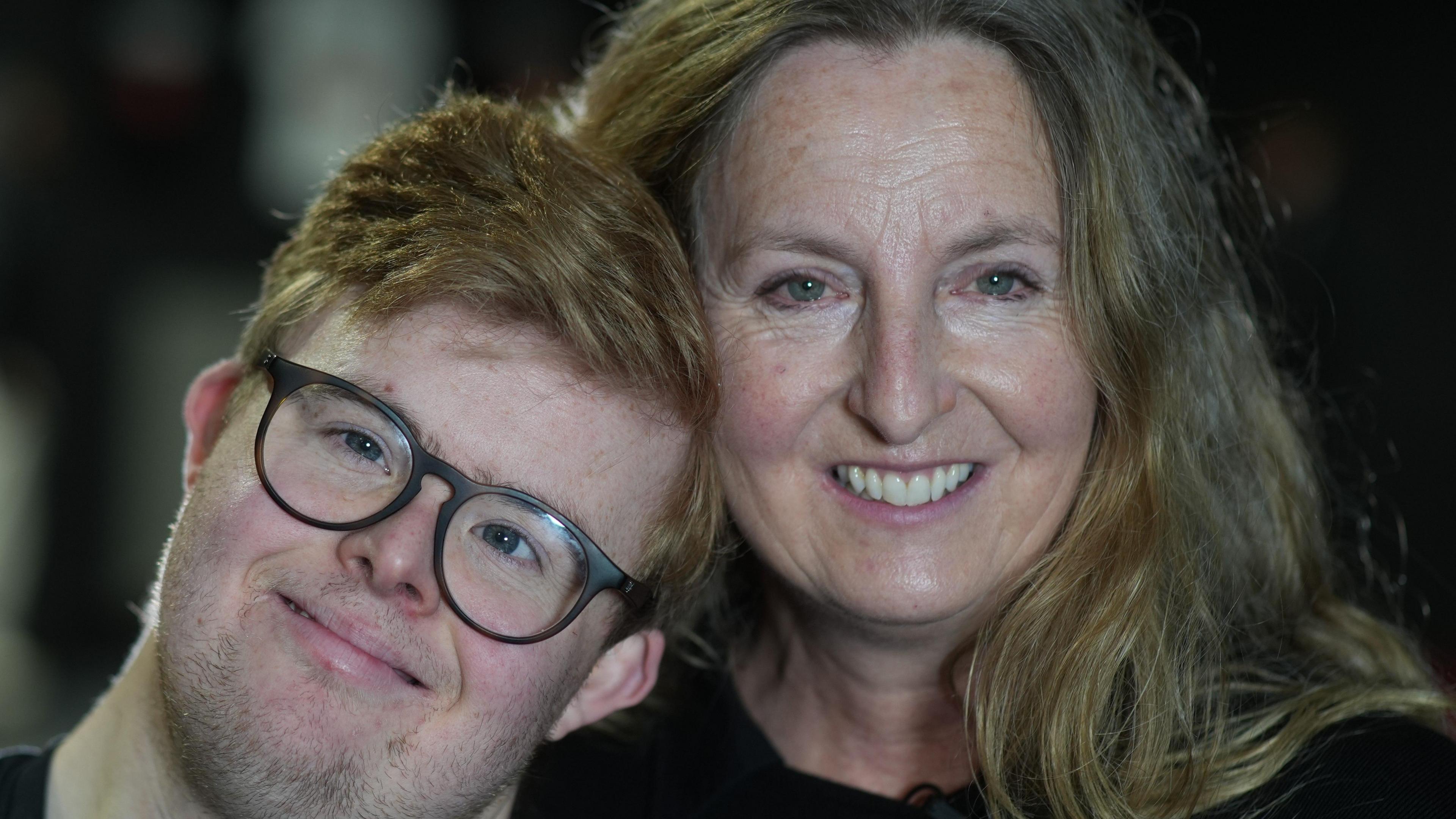 Edward Wagland with black glasses smiles at the camera while his head rests on a Helen Wagland's shoulder. She has shoulder length blonde hair with some grey. She is wearing a black top and smiling.