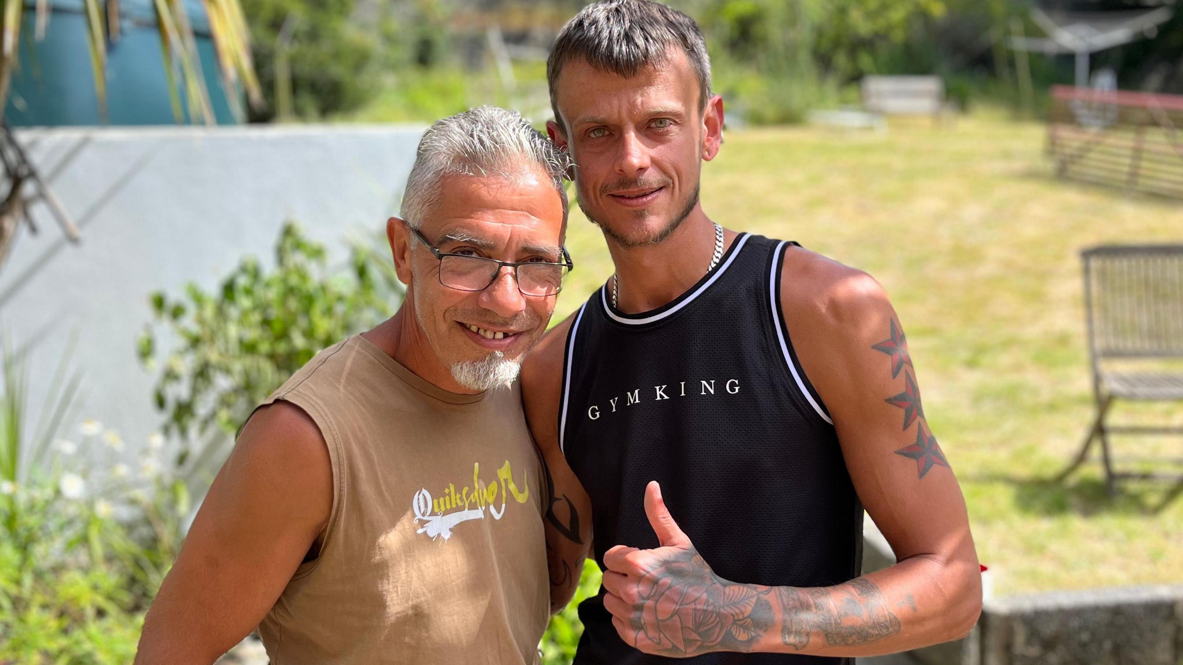 The two tanned men wearing sleevless t-shirts stand on a sunny day smiling at the camera with their arms around each other, one with thumbs up while the garden and garden furniture is slightly out of focus in the background.