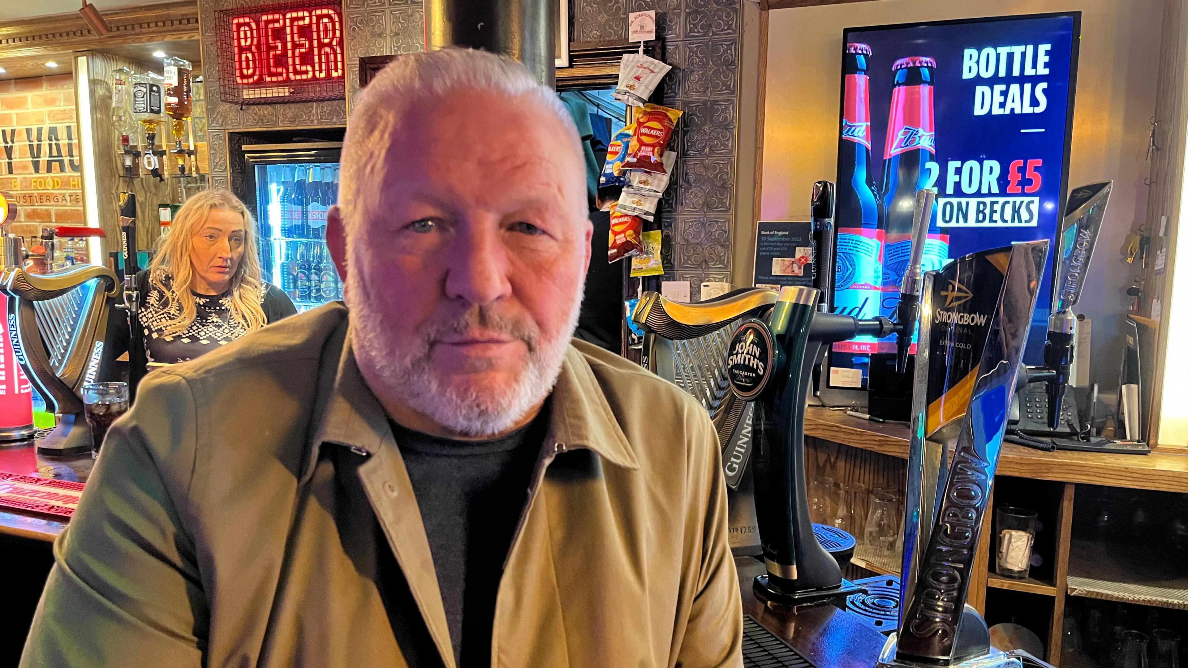 Man with white facial hair sits by a bar surrounded by beer pumps