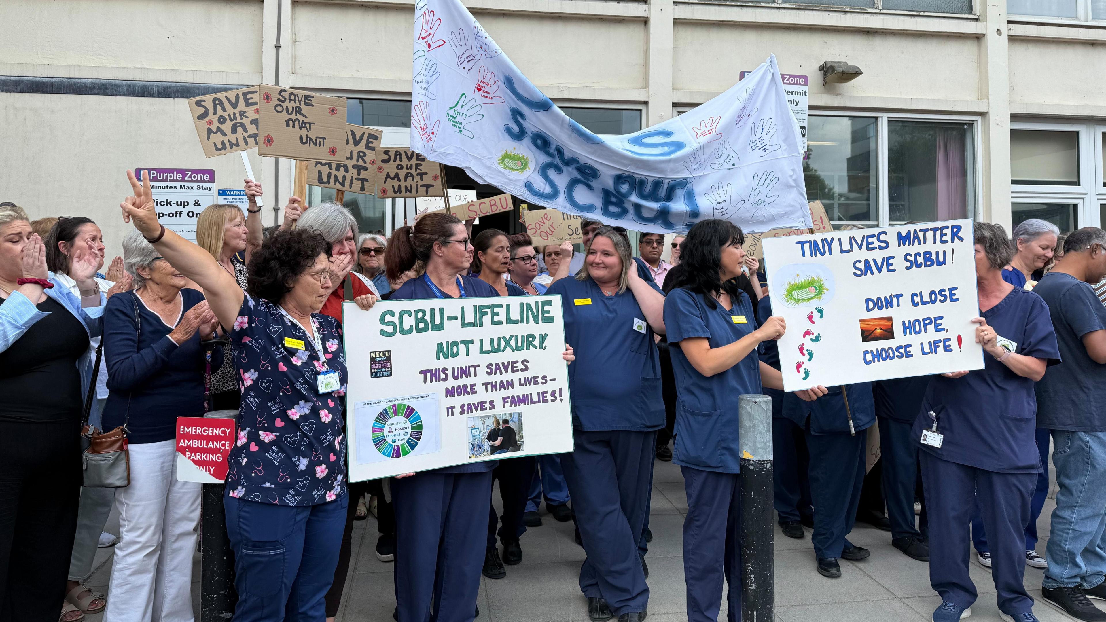 Nurses in navy scrubs and members of the public holding a protest outside a hospital building holding up home-made banners and signs reading things like 'tiny lives matter' and 'save our scbu'