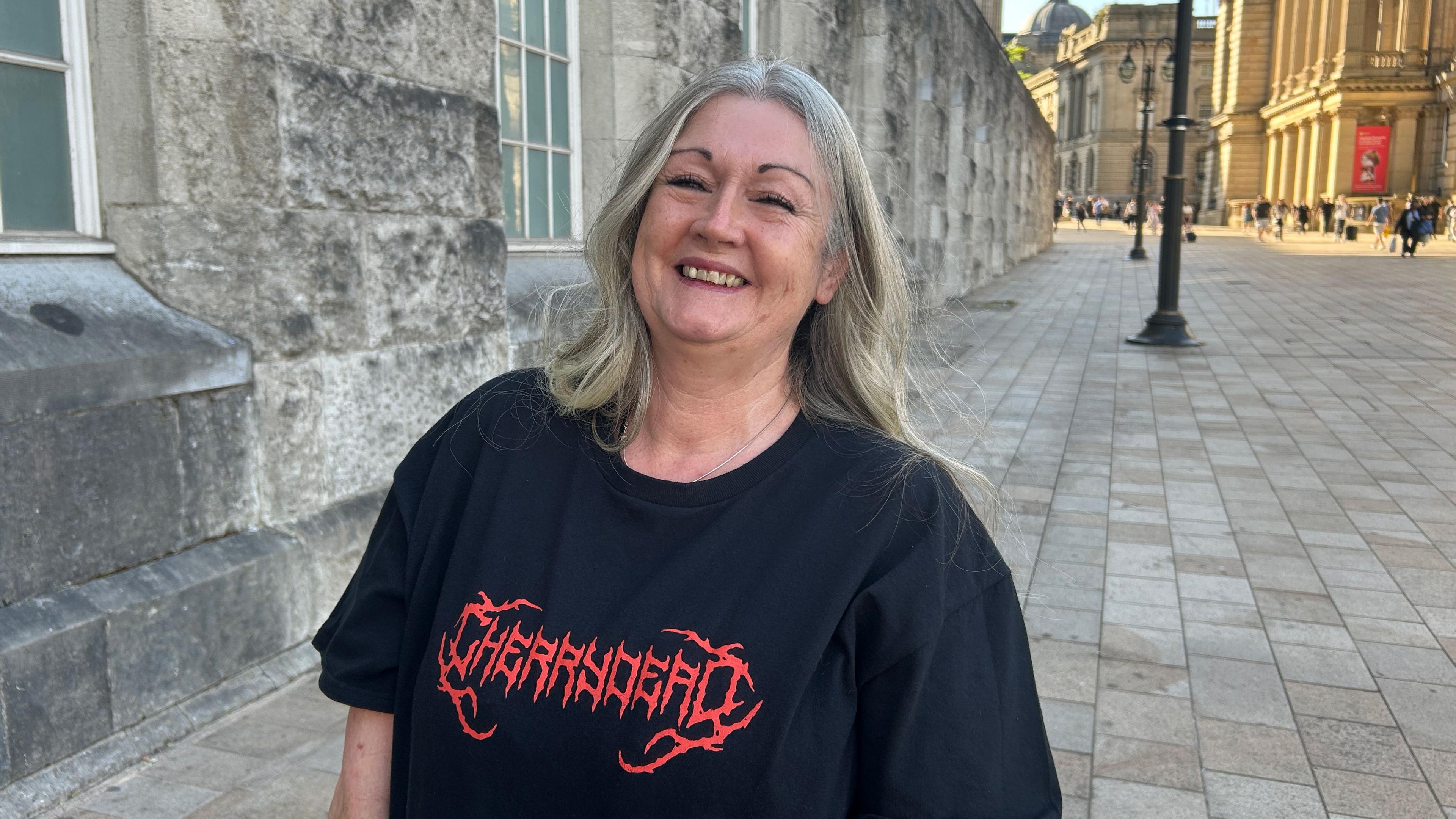 A woman with long hair in a black T-shirt with Cherrydead written in red letters on the front. She is stood outside on a brick pavement.