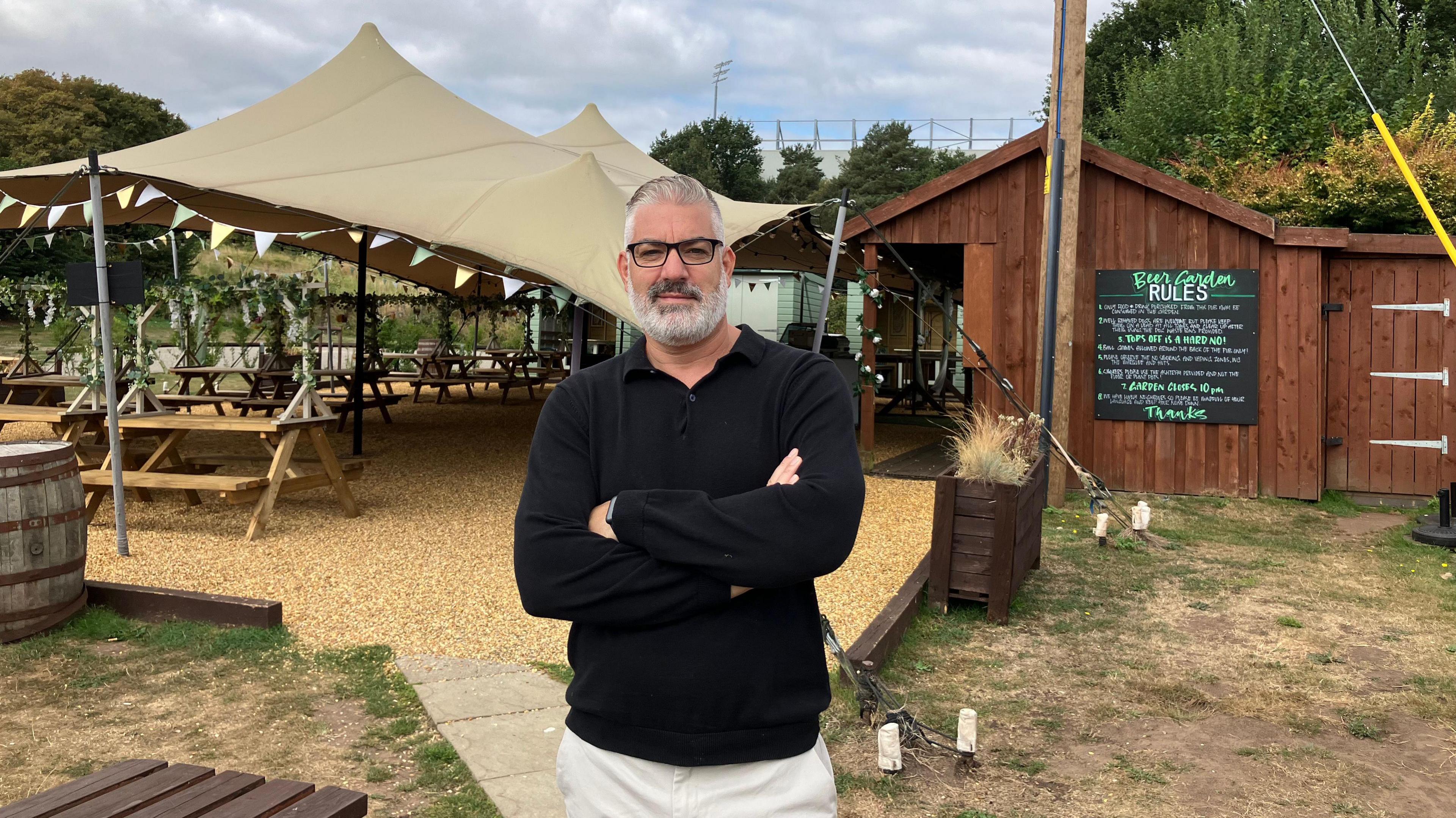 A man in glasses with grey hair and beard stands in front of the beer garden of a pub, with a large canopy and seats.