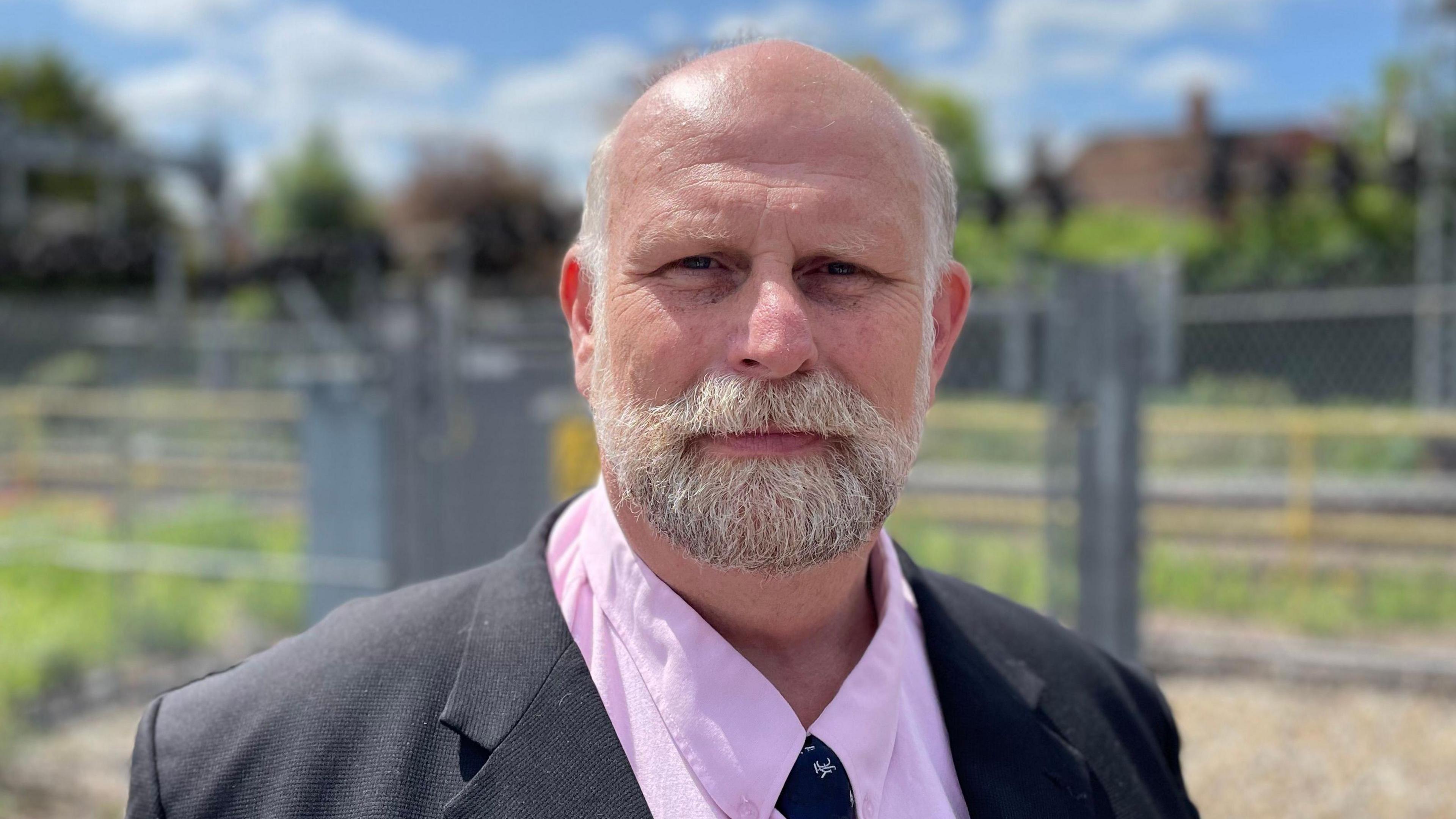 Tom Wotton with short white hair and a beard, looking at the camera, wearing a dark grey jacket, pink shirt and dark-coloured tie. He is standing in front of metal railings. There are grass and hedges beyond.