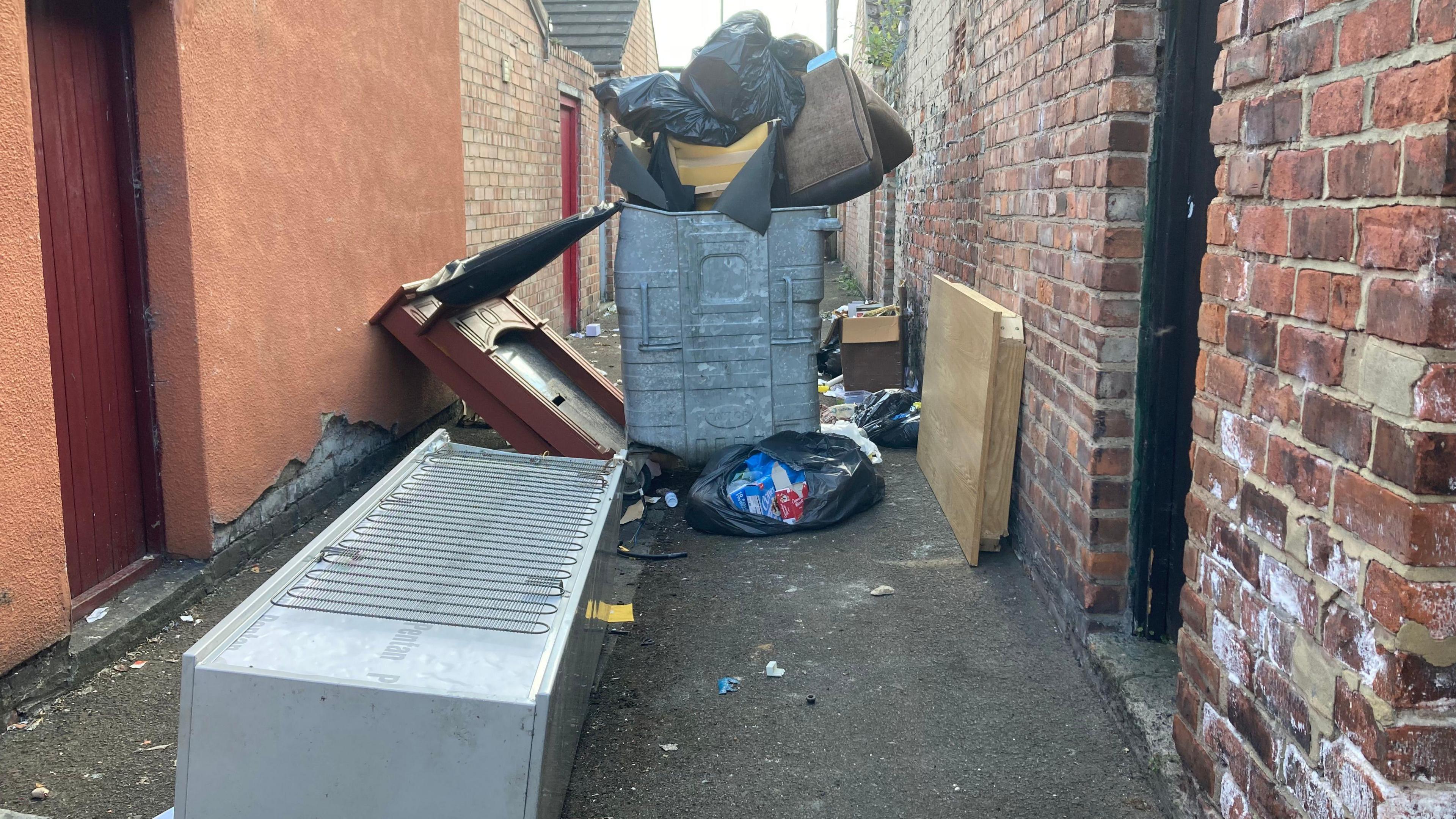 A large overflowing bin, a fireplace, and a fridge in an alley.