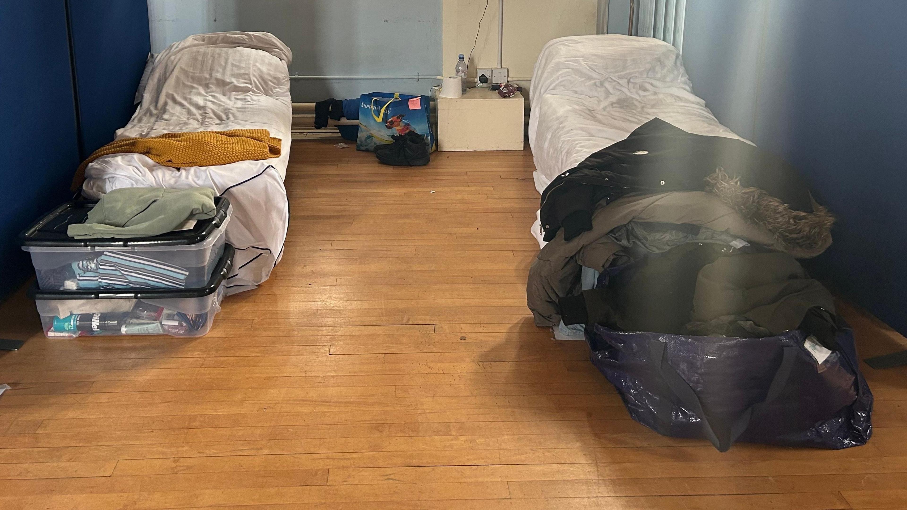 Two beds made up in a room, with boxes and bags at the bottom of the bed. The floor is wooden, and the walls blue. 