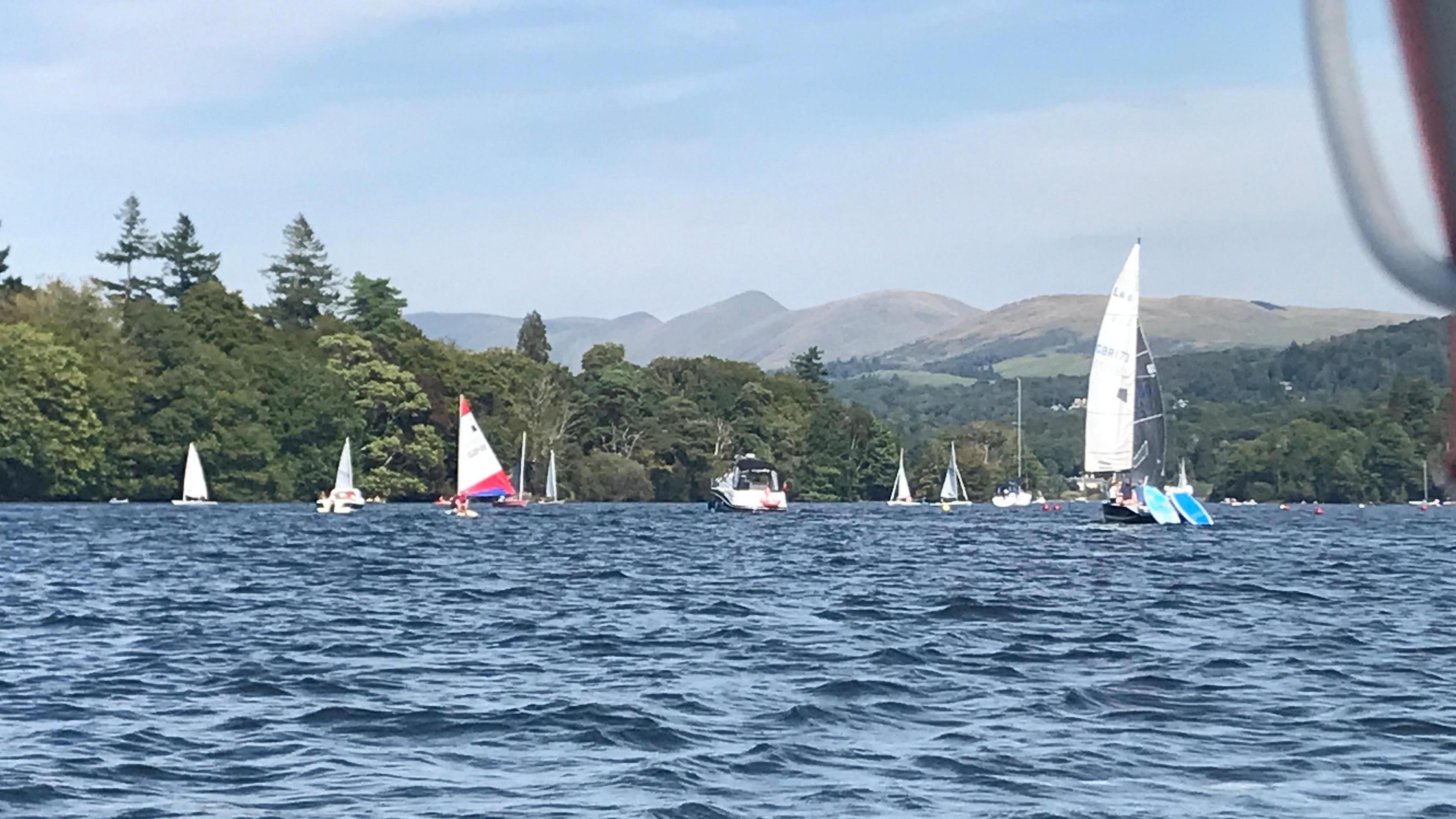 Sailboats on Bowness-on-Windermere. There are about seven or eight boats with all different coloured sails. There are hills in the background as well as trees.