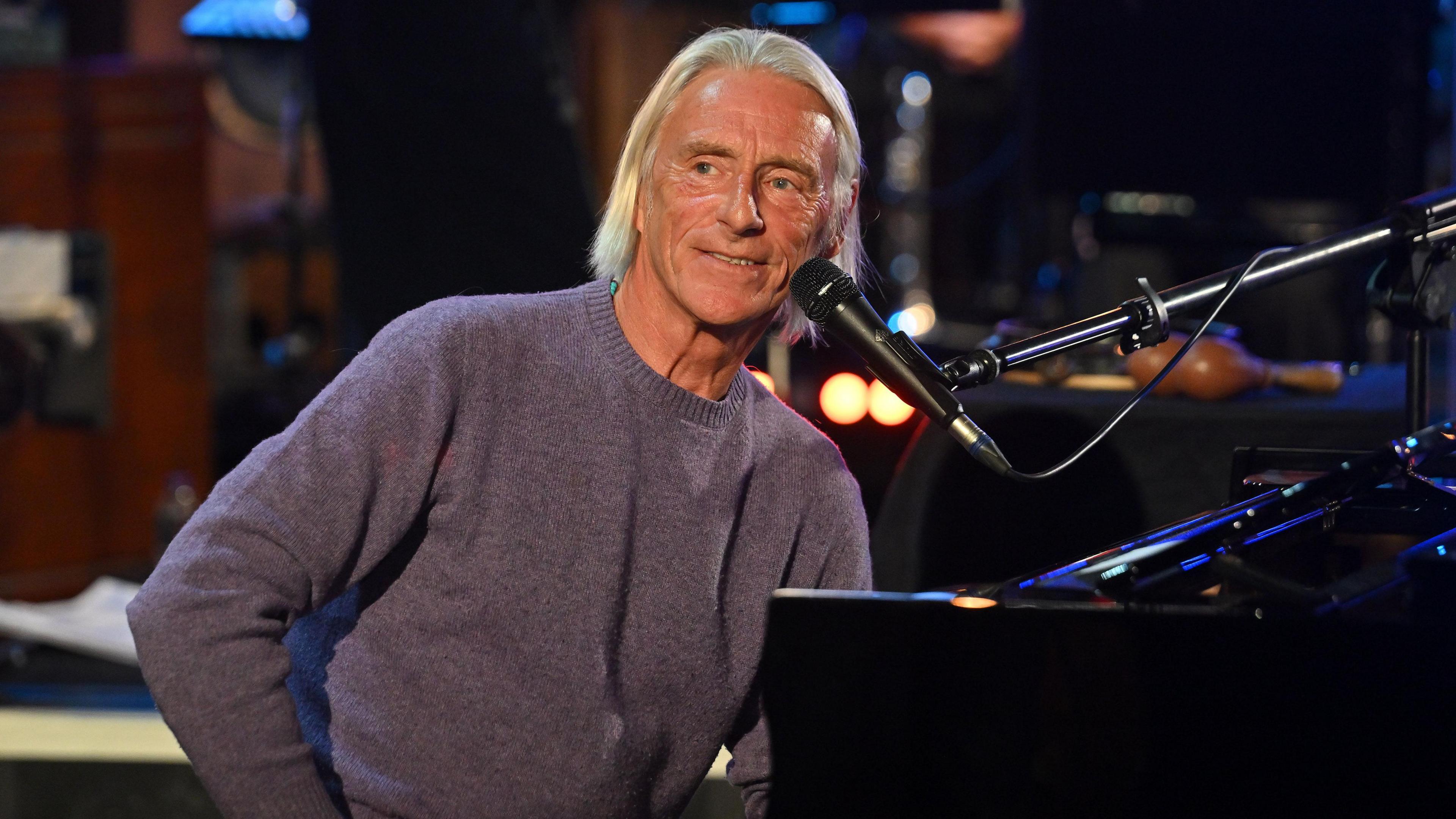 Paul Weller is sat at a piano, smiling. He has chin length blond hair and is wearing a purple jumper.