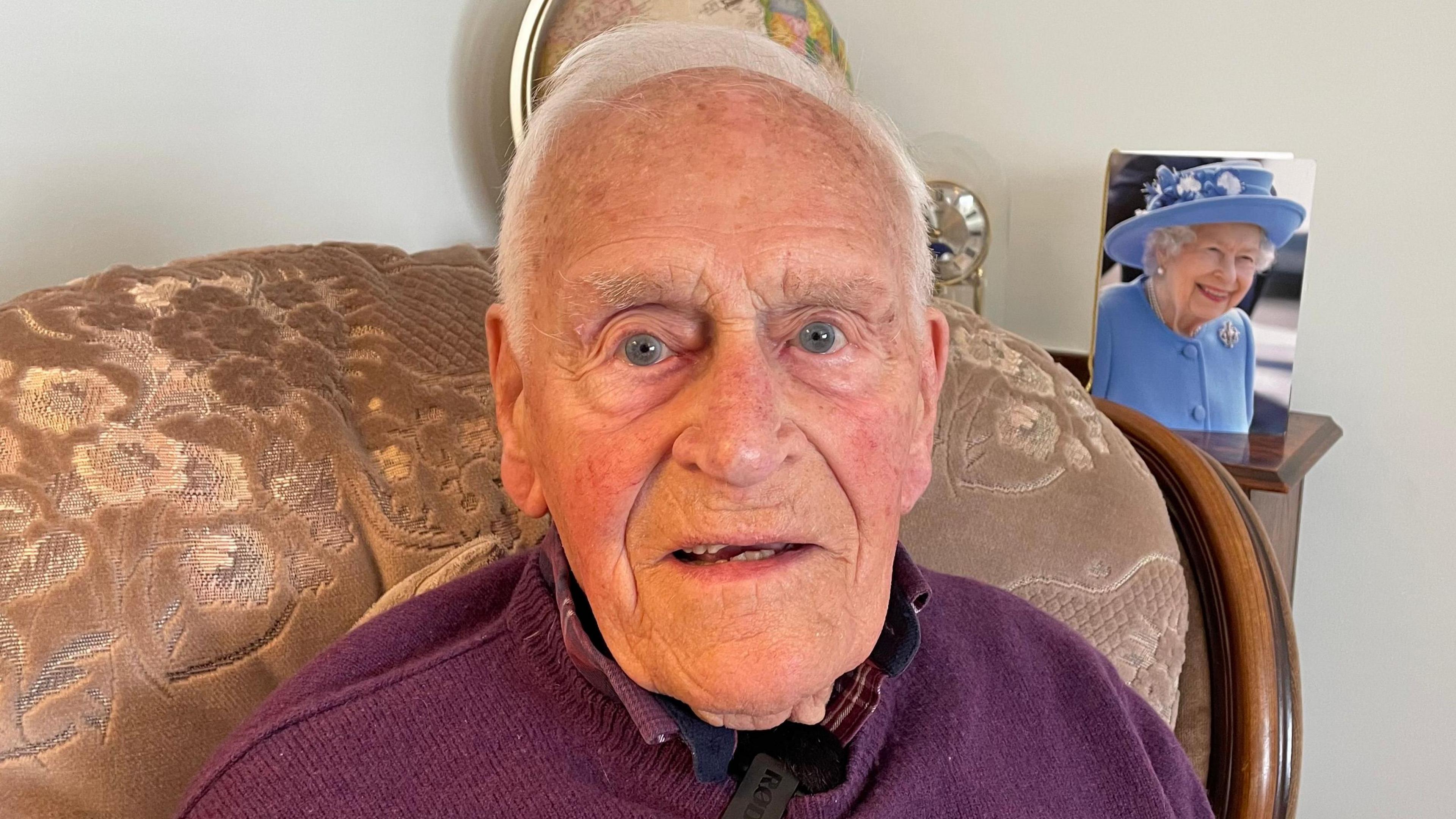 Monty Major sits in his armchair in his front room. He is an elderly man with short white hair. A card with a photograph of Queen Elizabeth, which he received for his 100th birthday, sits on a shelf behind him.