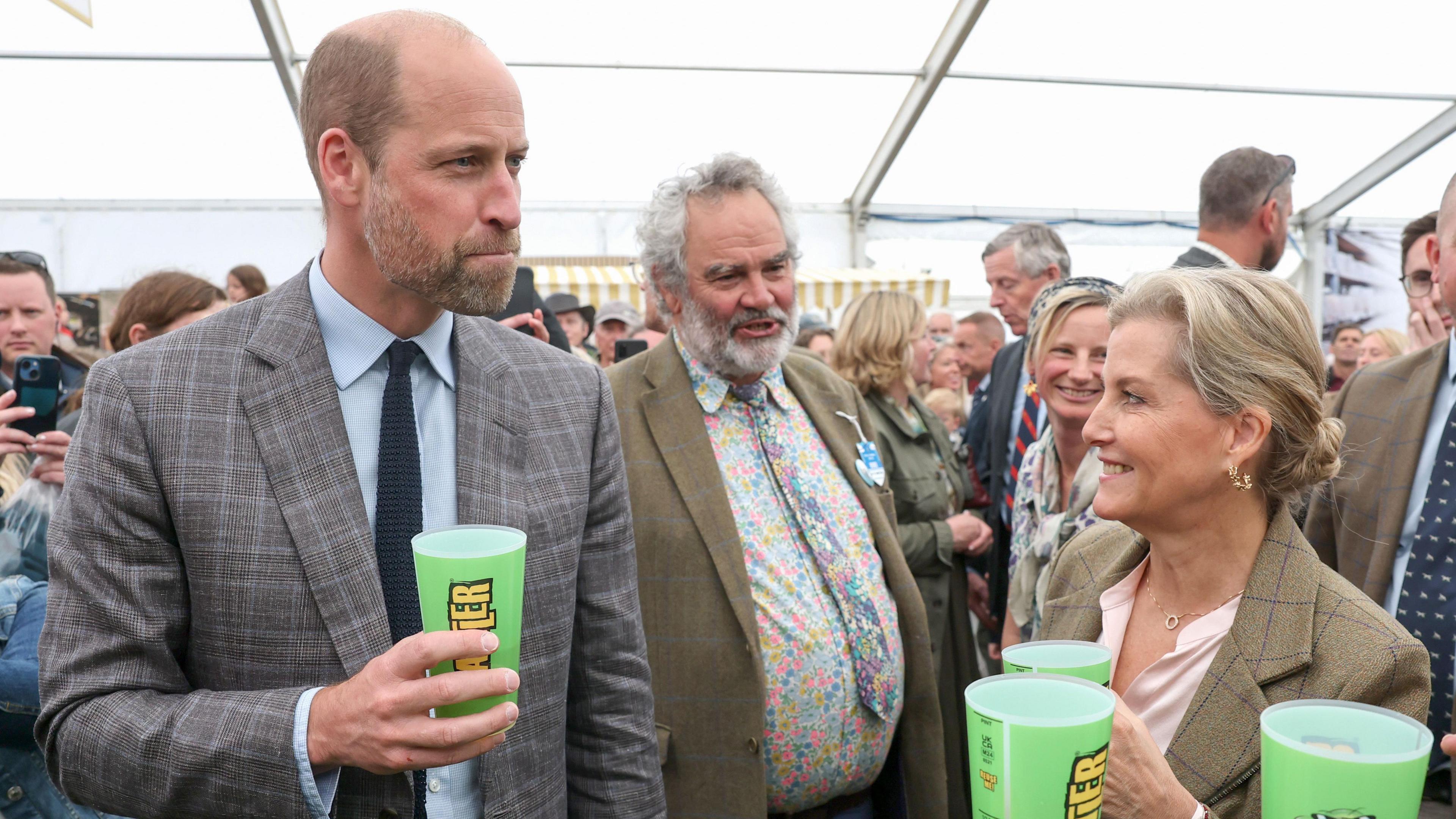 The Prince of Wales at the Royal Cornwall Show with his aunt Sophie, Duchess of Edinburgh. They are standing inside a tent and is surrounded by visitors to the show. They are both trying Rattler cider from a green plastic cup.