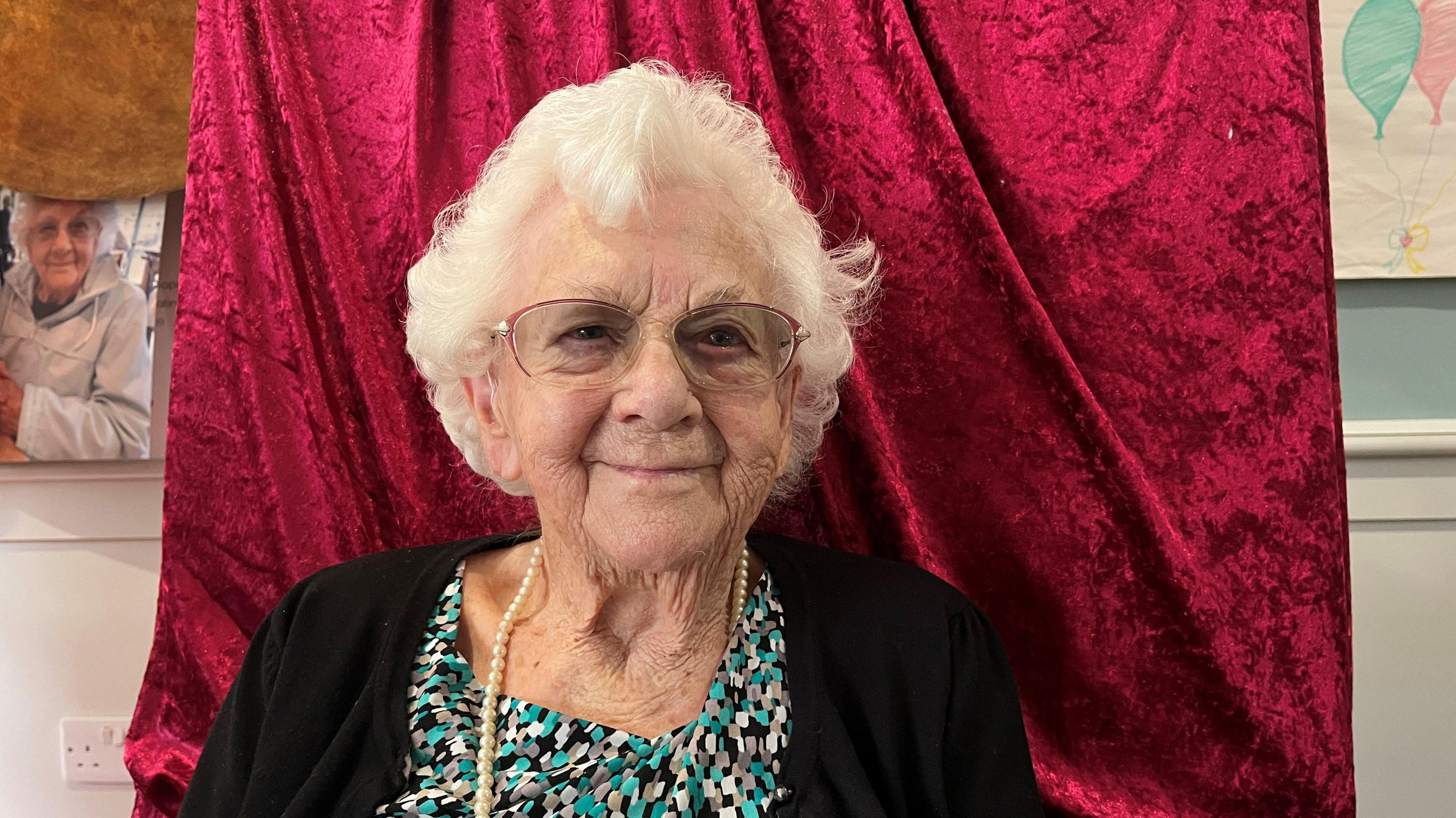 A head and shoulders shot of Sylvia Dalton who is sitting in front of a red curtain and wearing a dress with a string of pearls.