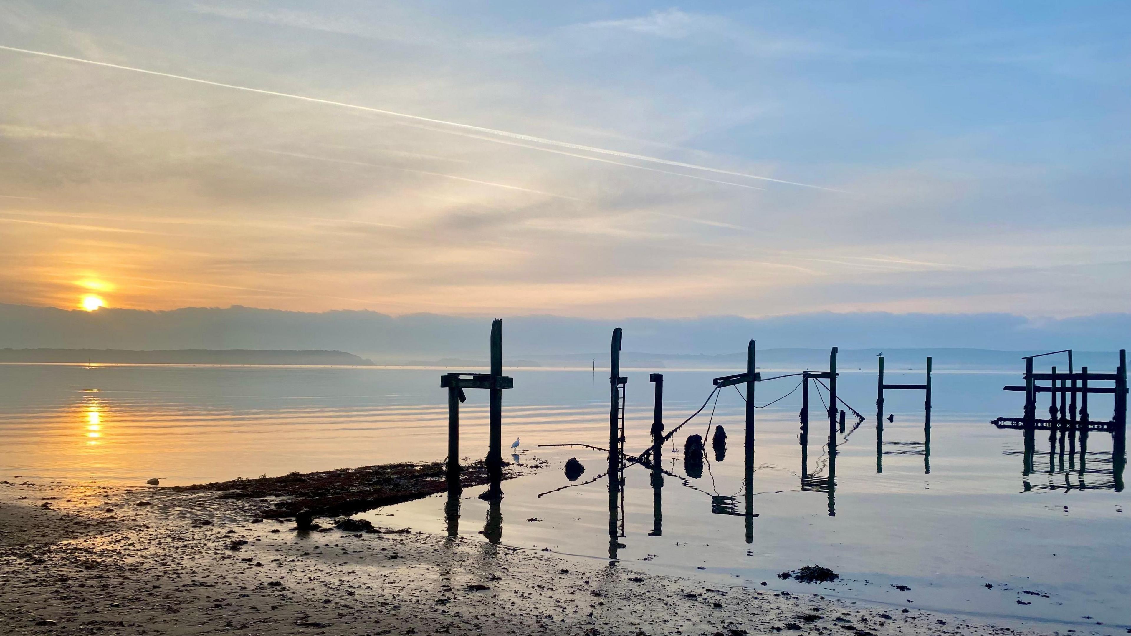 The sun is rising over the water at Hamworthy. There are gentle ripples in the water and hazy cloud in the sky. In the foreground you can see the beach and there are pieces of wood jutting out of the water.