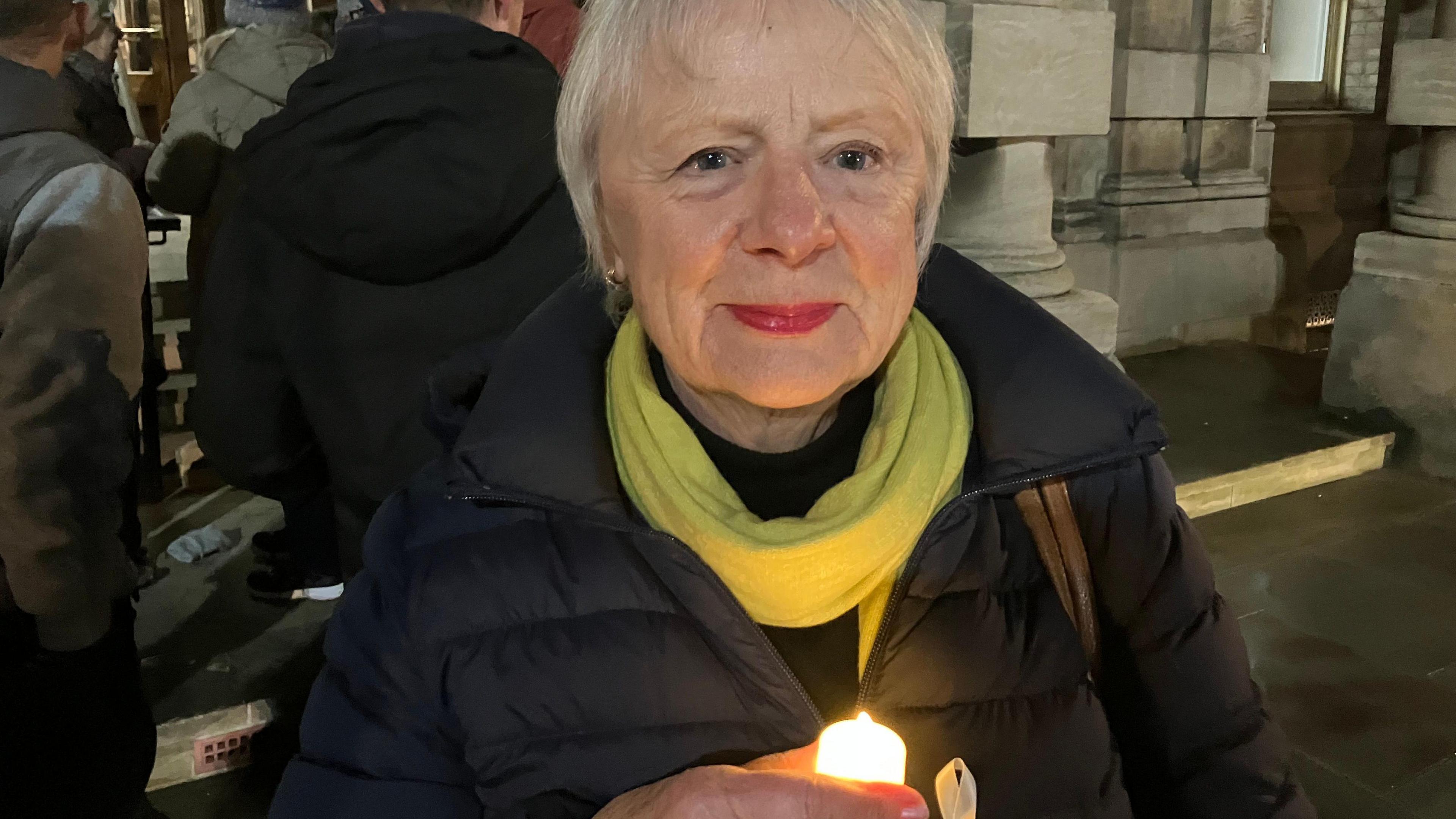 Woman with short white hair, green scarf and black jacket. She is holding a candle. She is stood on a street.