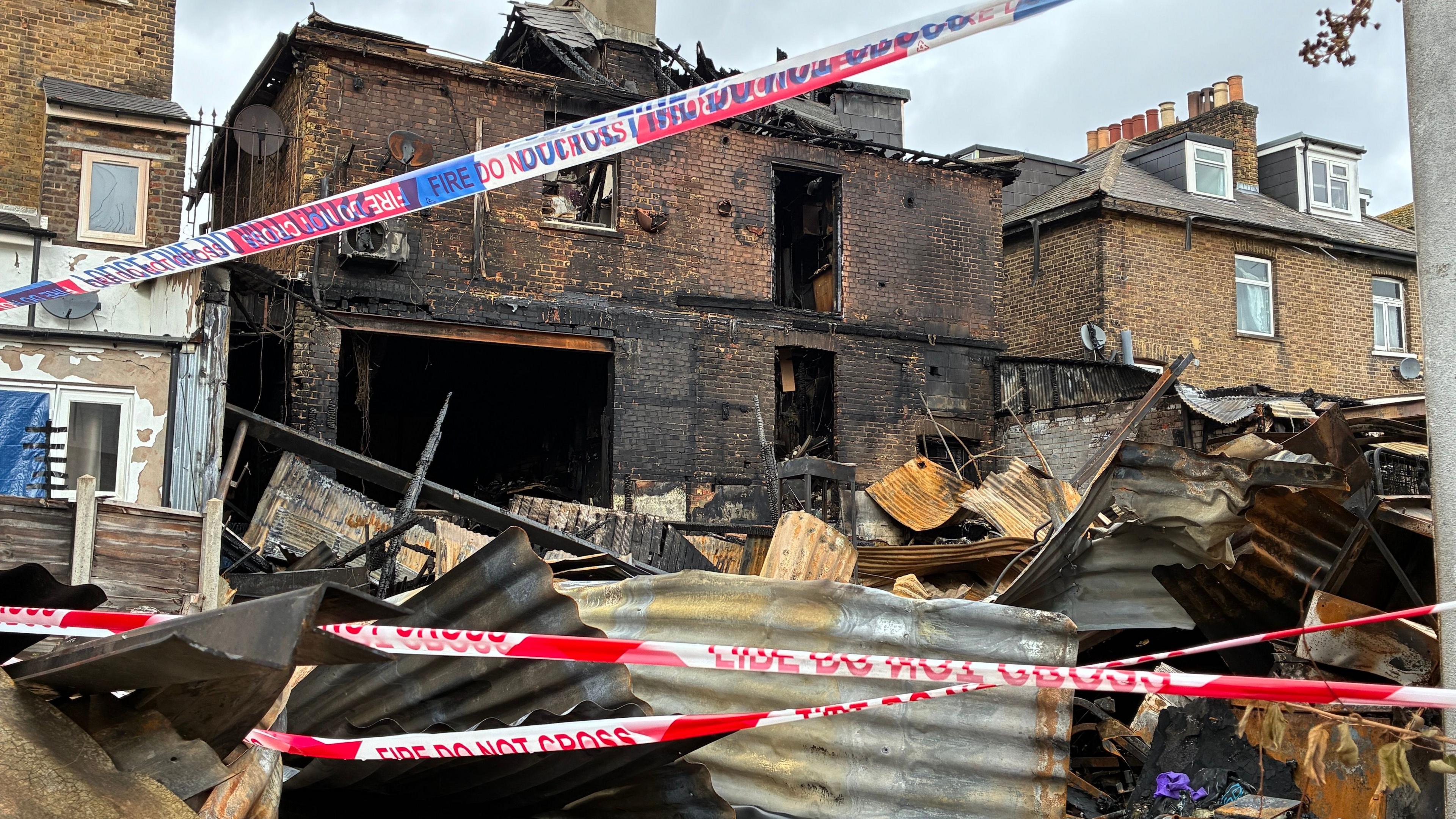 Police and fire brigade tape crosses an area of charred corrugated metal, the badly burned brick building in the background