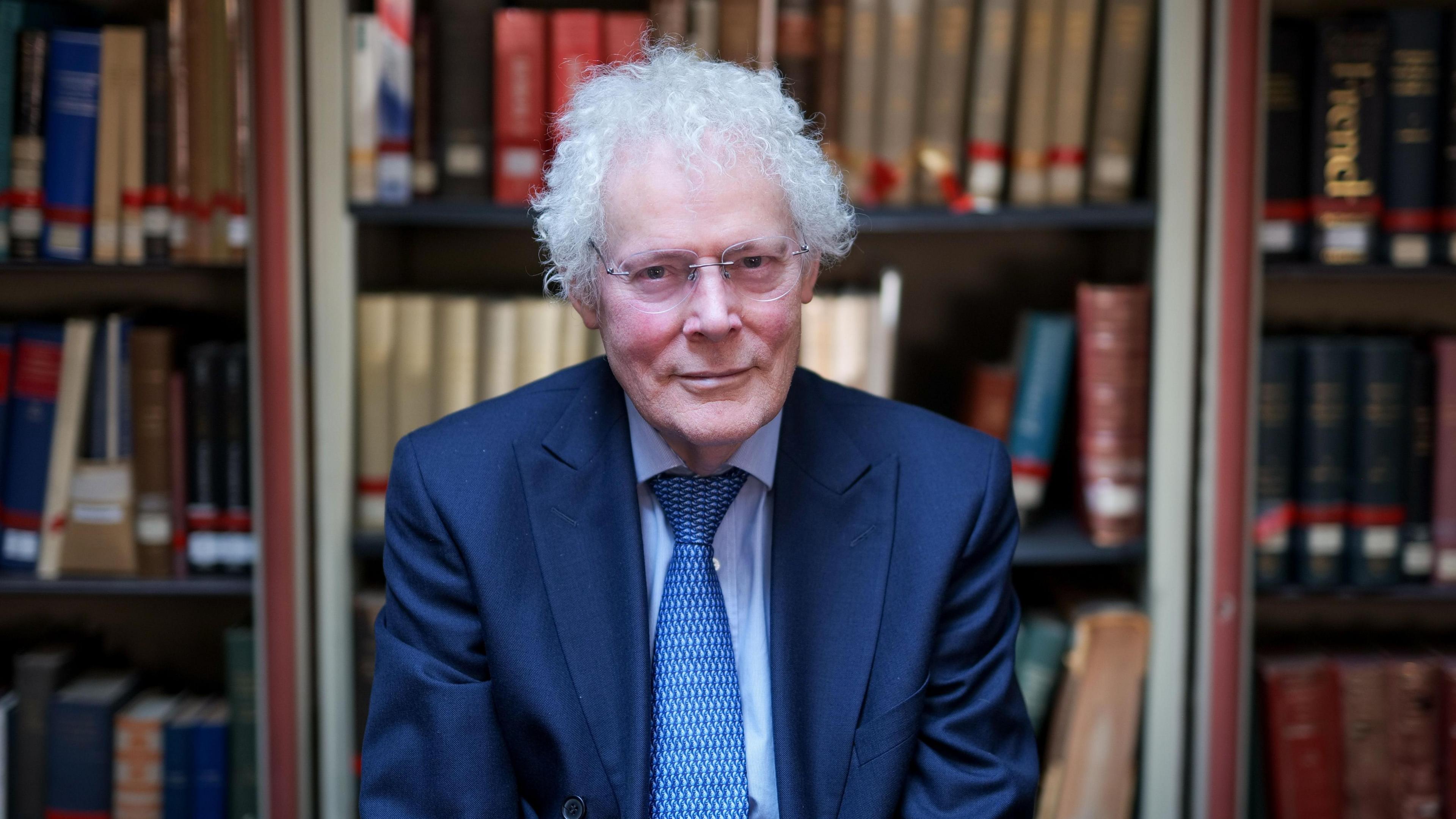 Professor David Carpenter from Kings College London wears glasses, a blue suit and tie. He sits in a library looking at the camera in front of bookshelf.