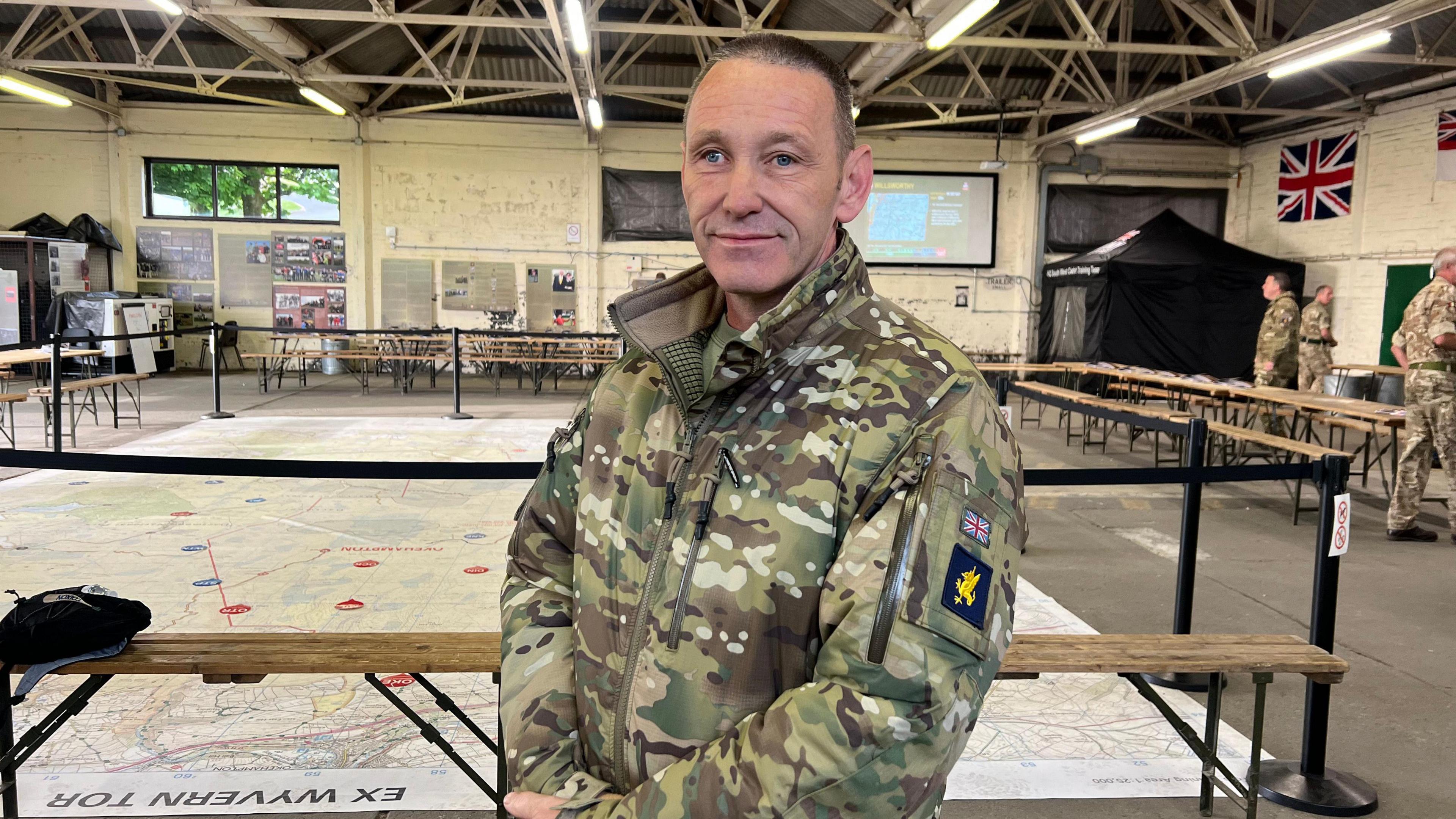 Sgt Tosh Hodgkins stood in the control centre at Okehampton Camp on Dartmoor. There is a big map on the floor behind him and other military personnel in the background. He is wearing camouflage military clothing.