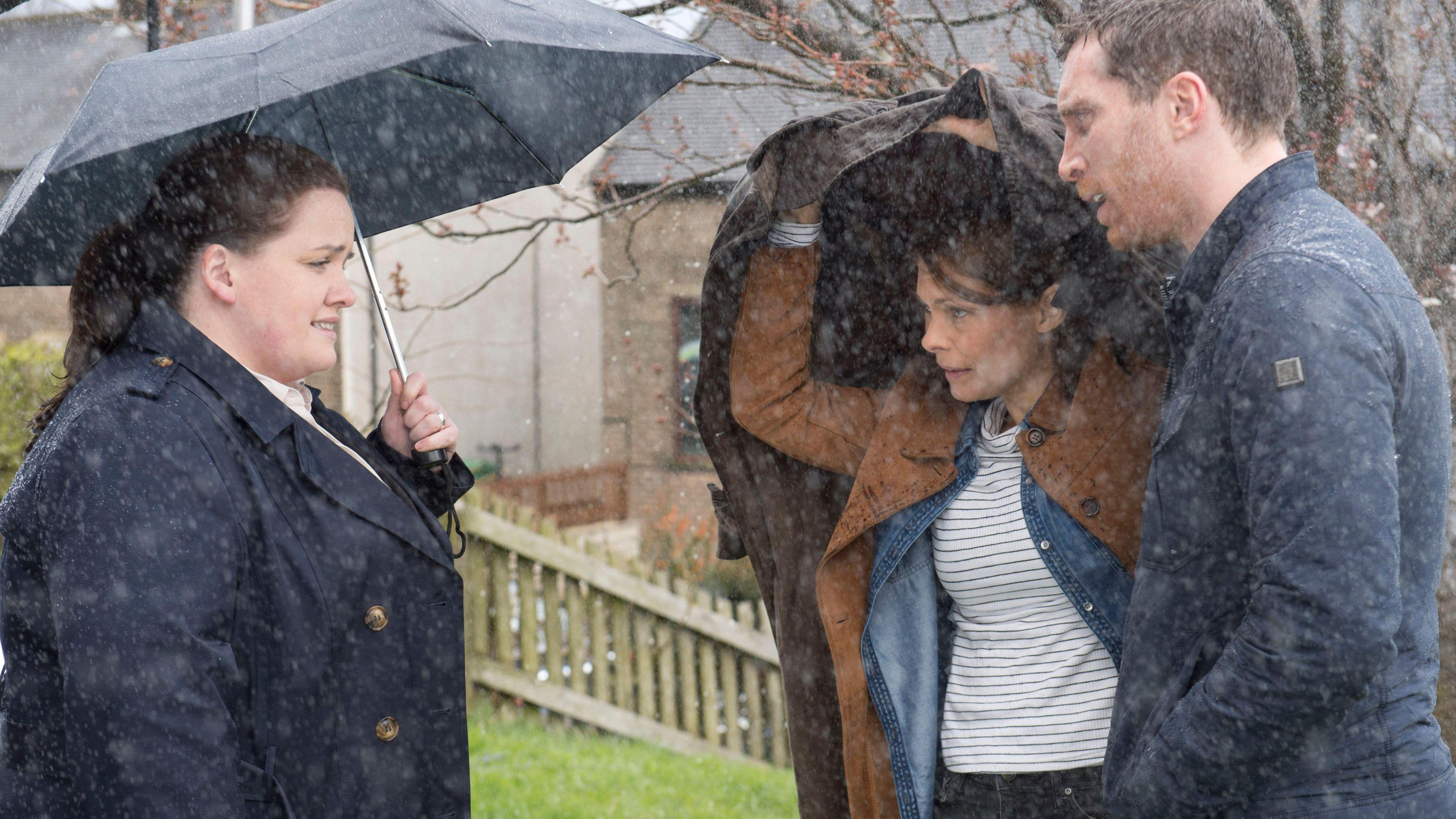 A woman stands under an umbrella talking to a man and a woman, the woman sheltering under a coat