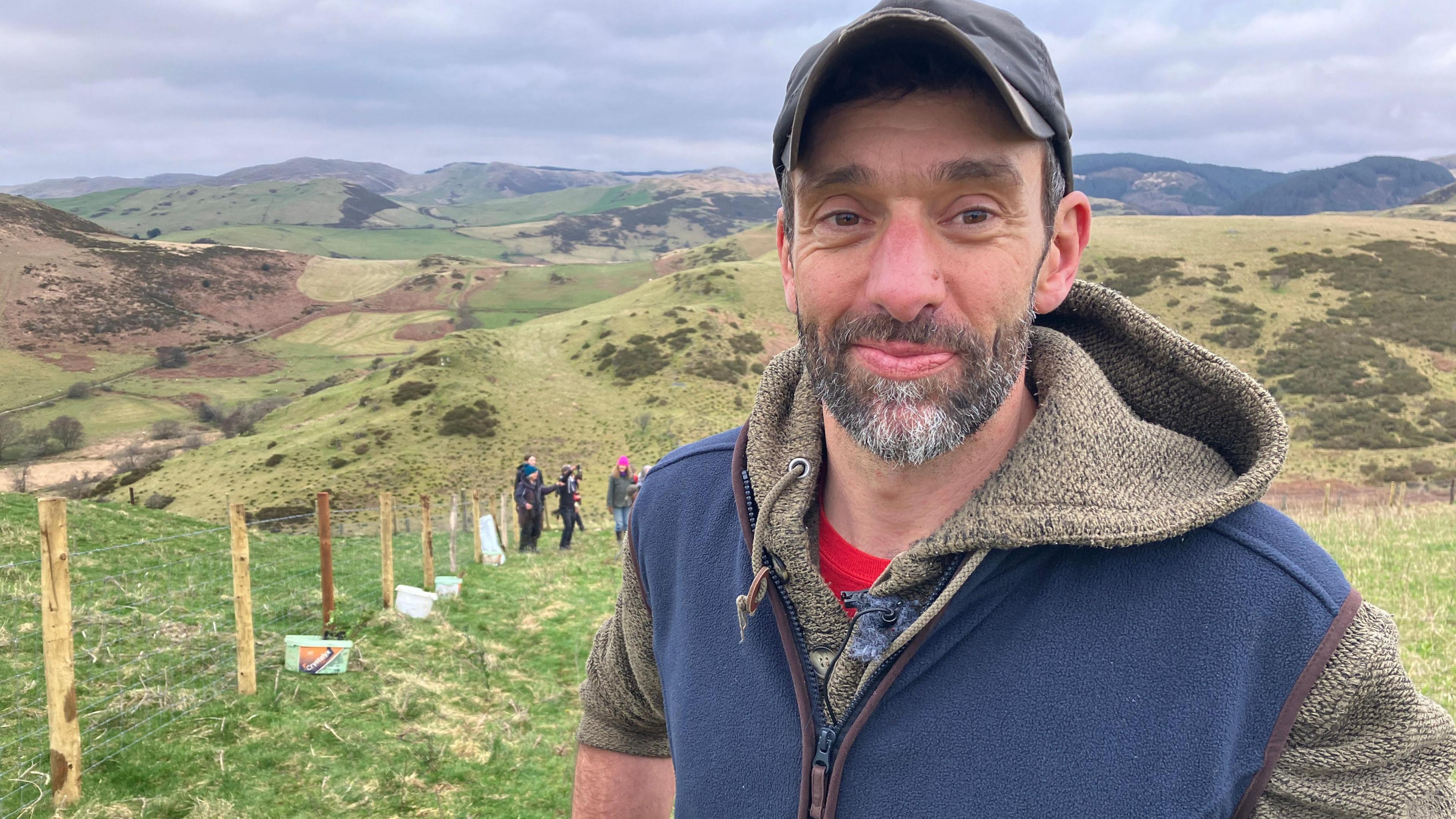 Llun o Rhodri Lloyd-Williams yn sefyll ar gae ar dir ei ferm - mae'n gwisgo cap tywyll a gellir gweld bryniau yn ymestyn i'r pellter y tu ol iddo. 