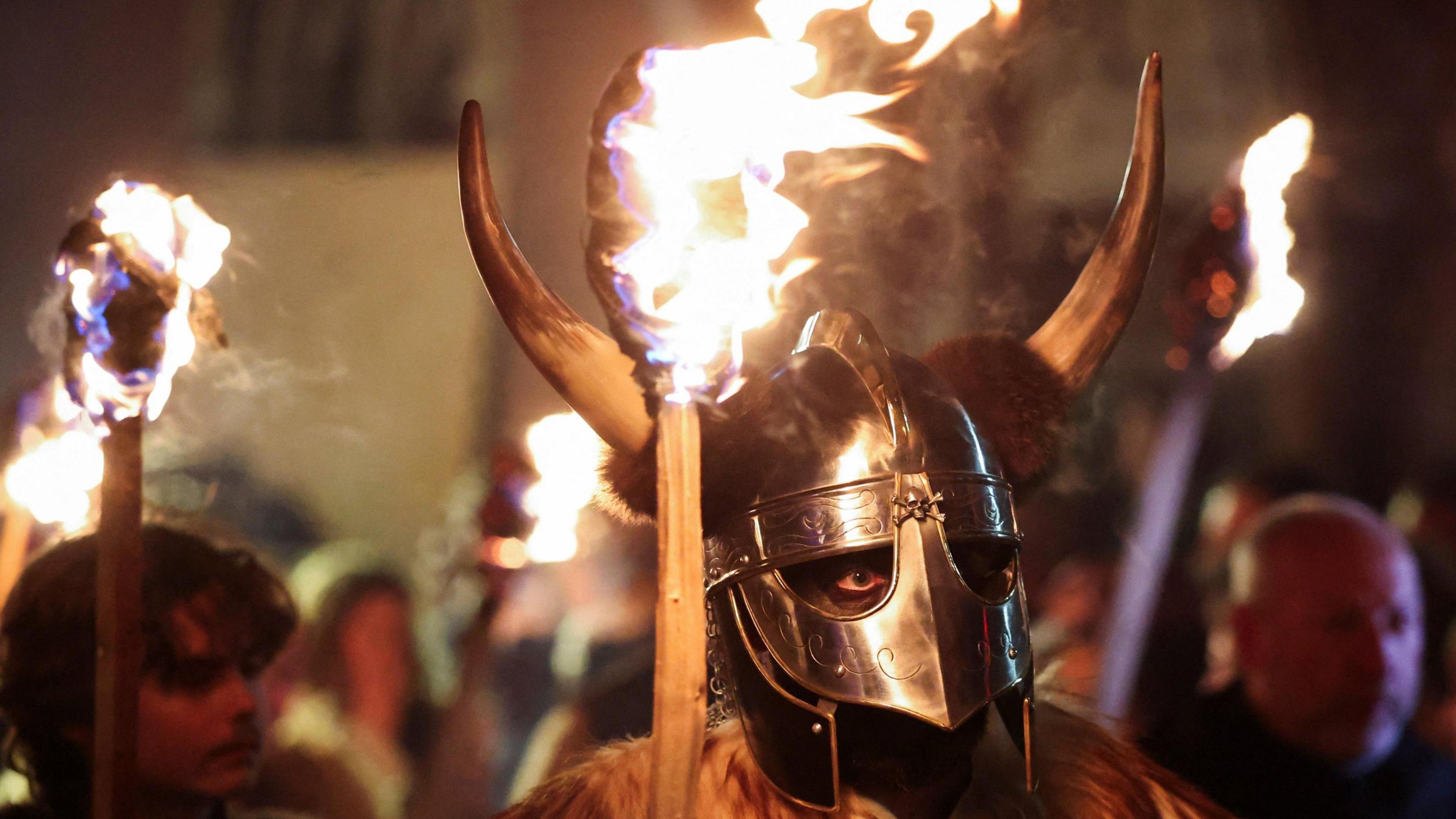 A man wearing a viking helmet, with mask and bull horns carries a burning torch.