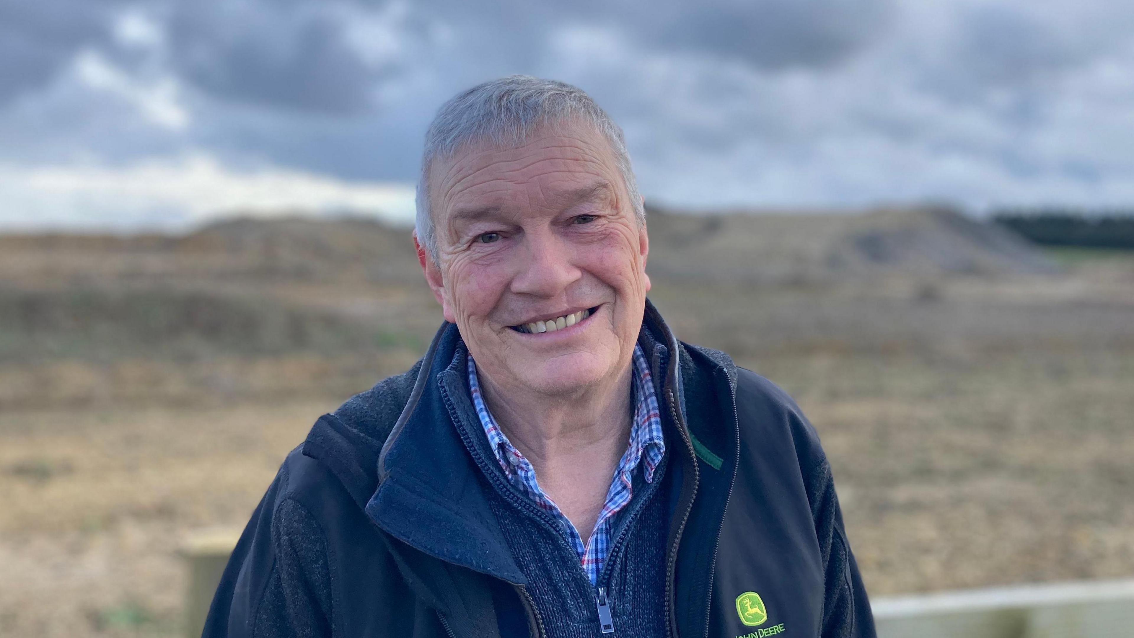 Stephen Adkins in a black fleece, blue jumper and checked shirt in front of a fence with a field behind