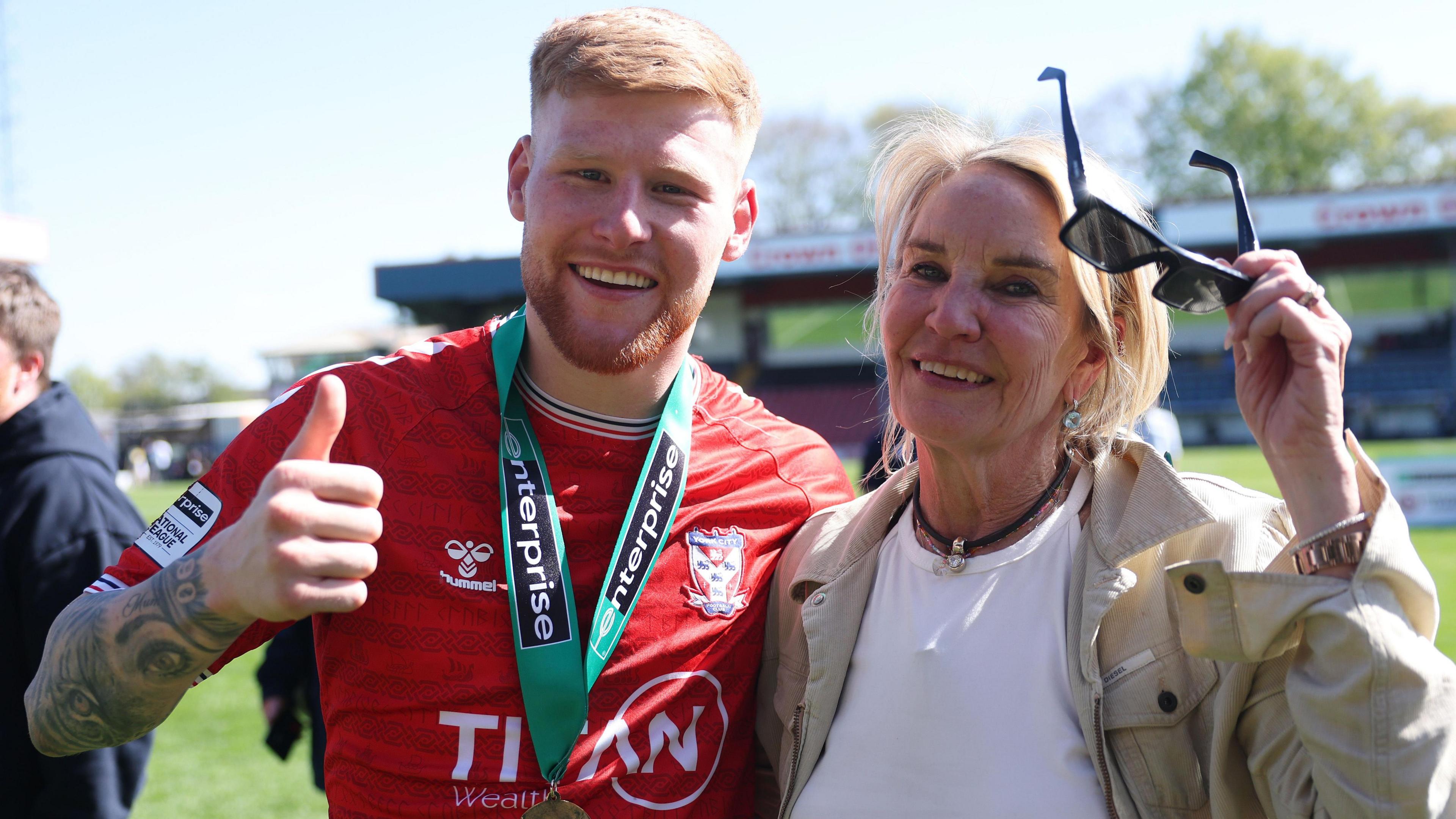 Josh Stones, wearing York City's red shirt with a National League title-winners medal around his neck, smiles and puts his thumbs up while holding his arm around York City owner Julie-Anne Uggla, who is smiling while wearing a white shirt and beige jacket.