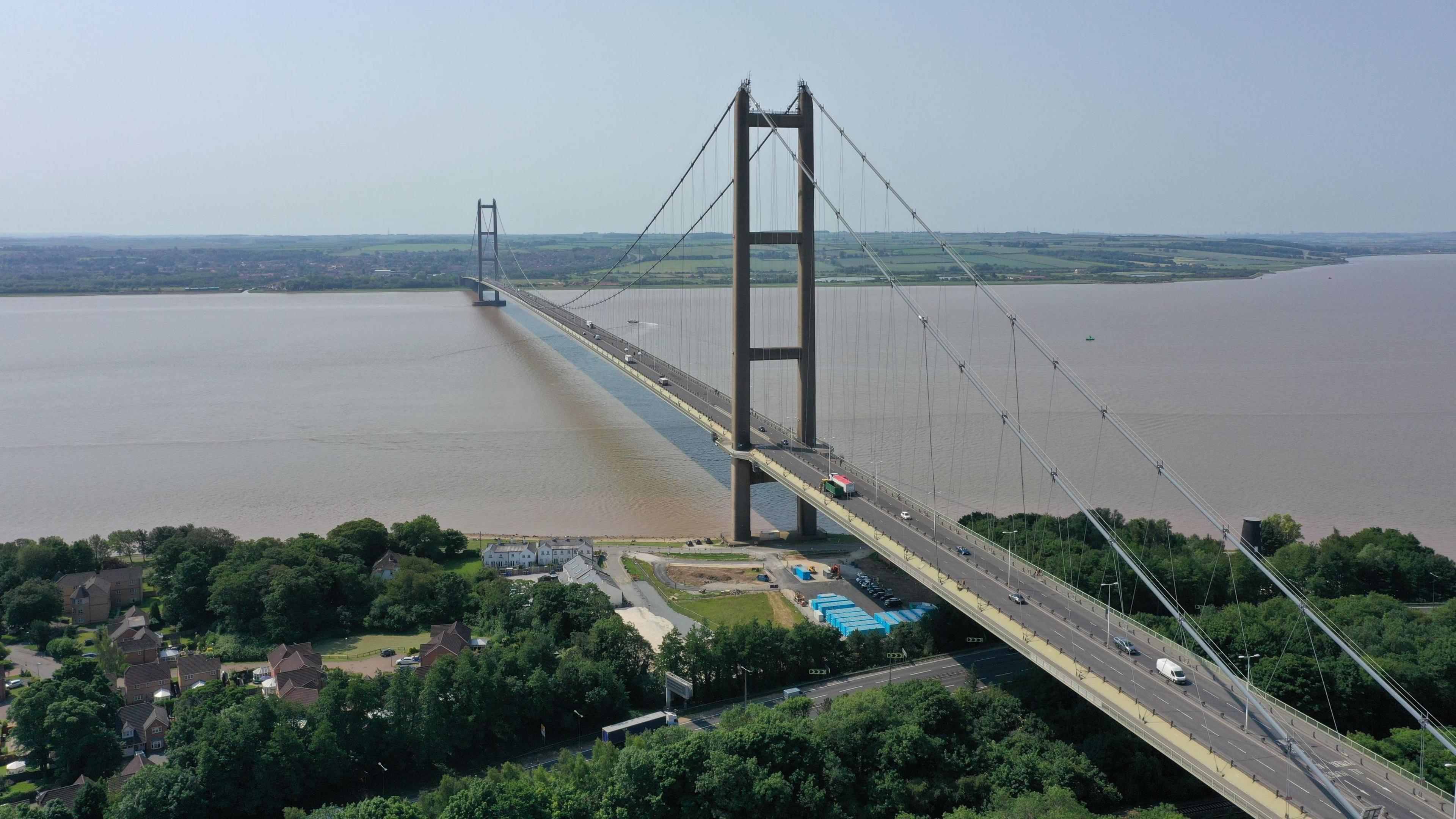 The Humber Bridge stretching across the Humber. The bridge has tall towers and cables supporting it, with vehicles traveling in both directions. Green countryside and scattered houses can be seen on either side of the Humber. The sky is clear and blue.