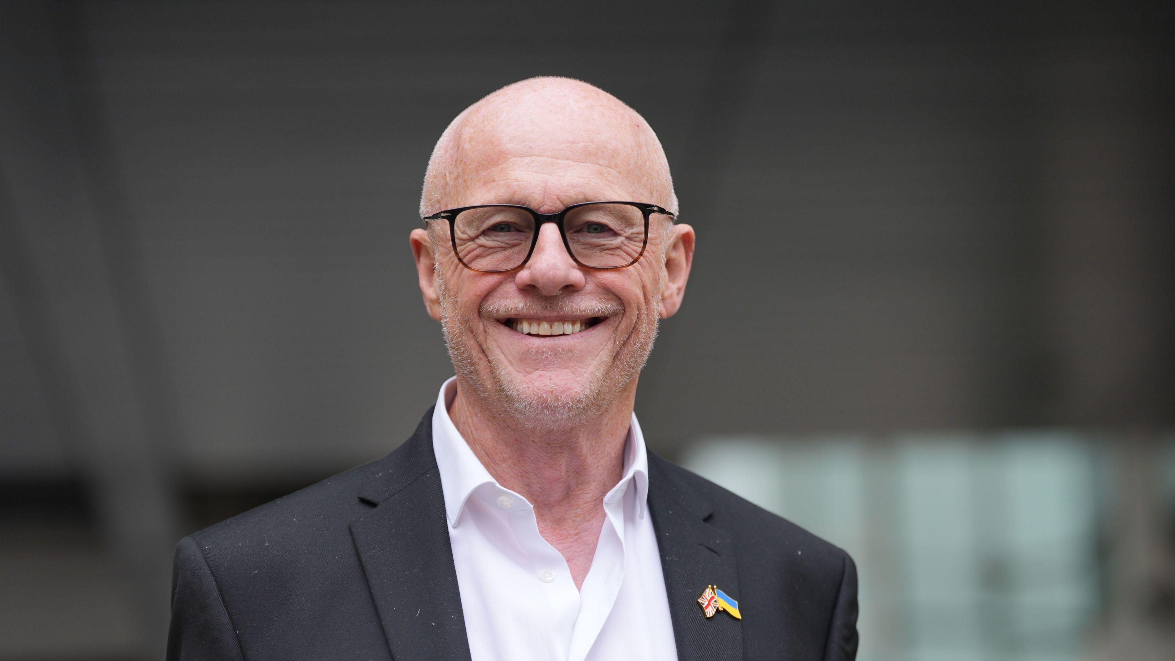 Businessman John Caudwell leaving BBC Broadcasting House in London - he is wearing a black dress jacket with a Union Flag and Ukraine flag pin on his lapel - he has a bald head, a broad smile and is wearing dark rimmed glasses, with his white shirt unbuttoned and with no tie 