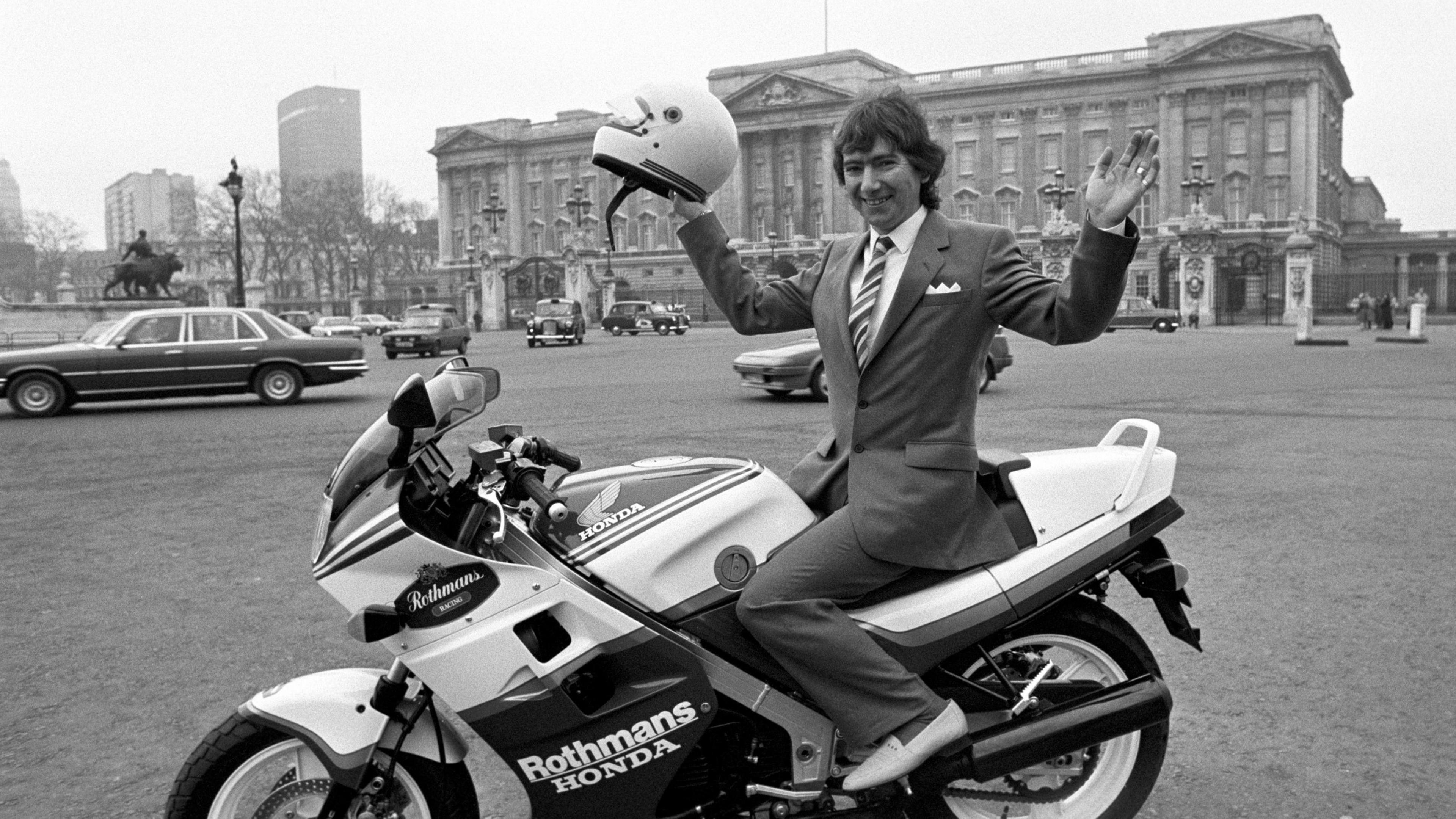 A black and white photo of Joey Dunlop, a man with wavy brown hair, he wears a suit, and has his arms up in a pose as he holds a white helmet and sits on a white motorbik e in front of Buckingham Palace.