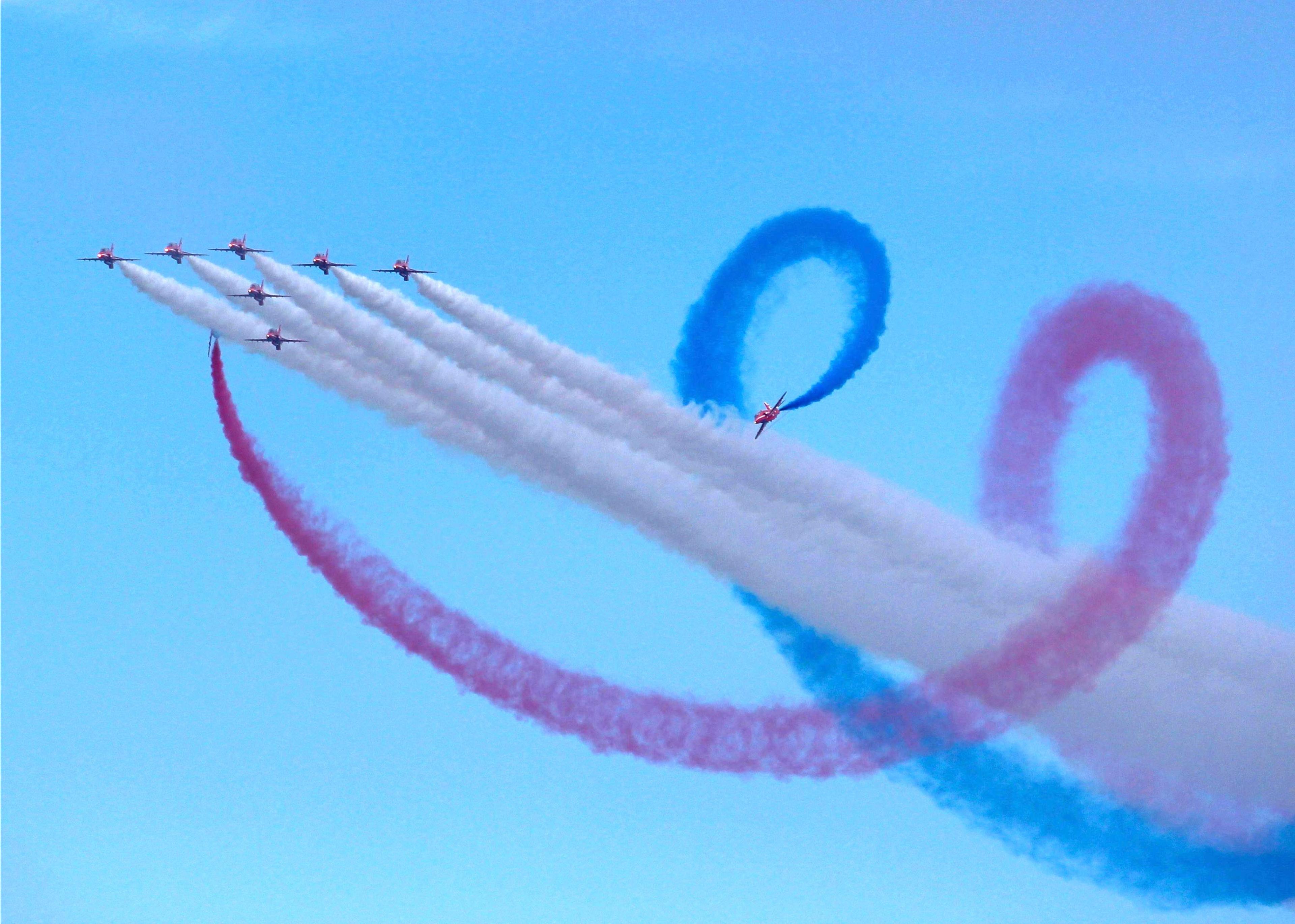 The red arrows performing - seven of them flying in a V-formation and three circling the white smoke coming from them while emitting red and blue smoke - against a bright blue clear sky