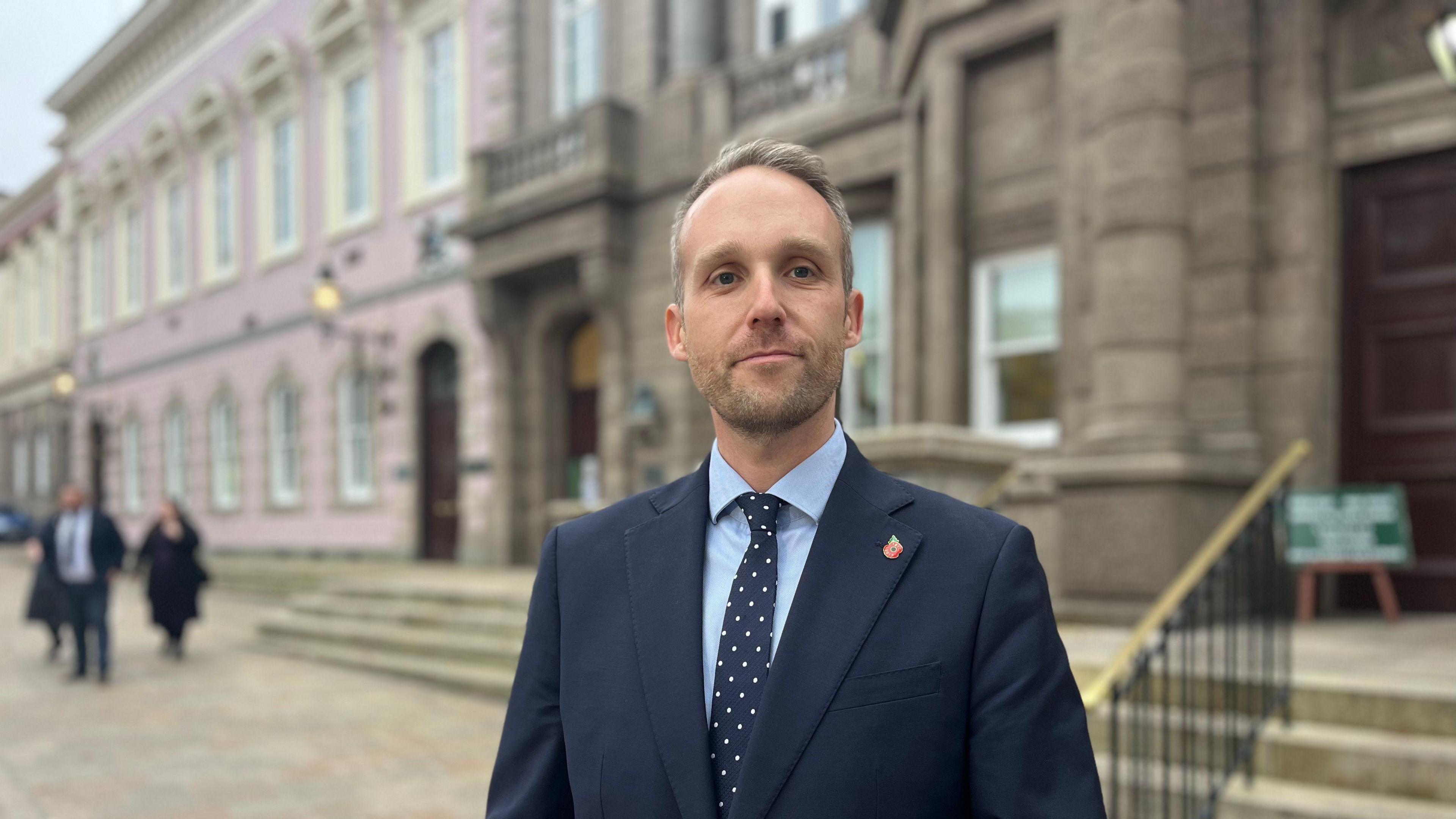 Det Insp Aiden Quenault pictured outside Royal Court wearing a navy blue suit, a match tie with white polka dots and a light blue shirt. He has mousy brown hair that is swept to one side and light stubble.