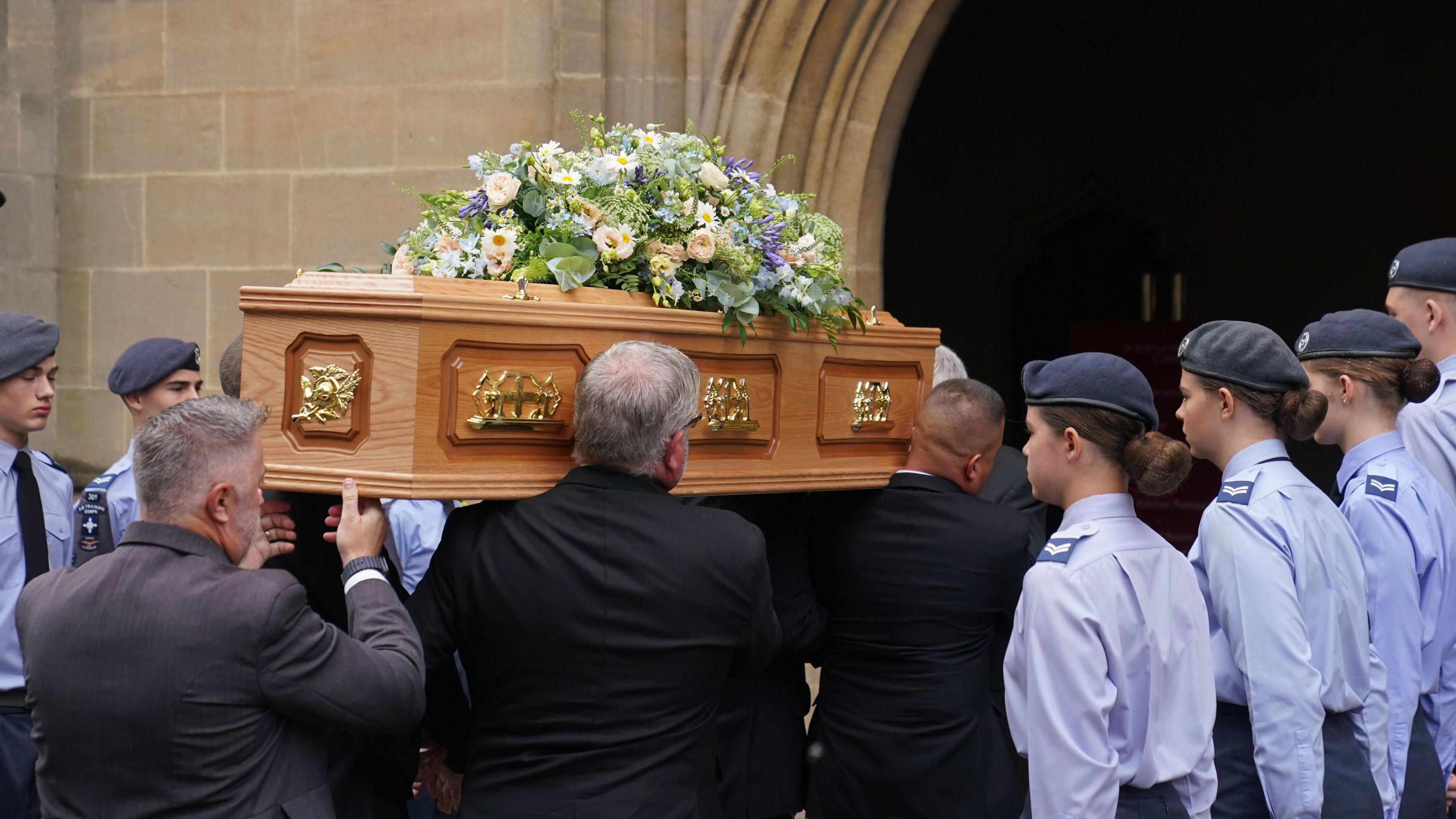 A light brown wooden coffin is carried on shoulders inside a doorway, young people in light blue shirts wearing berets stand guard either side of the coffin.