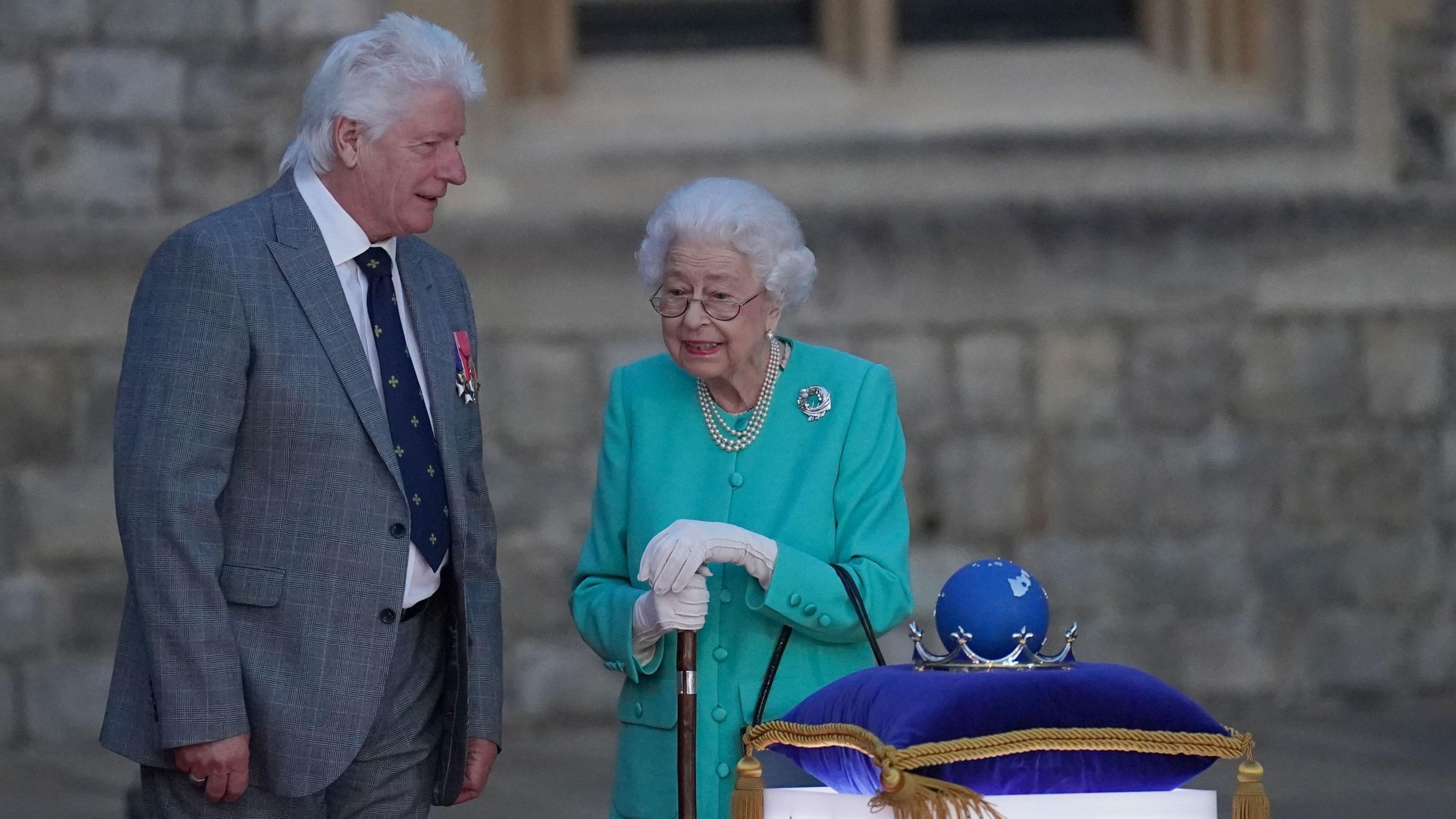 The Queen wears a turquoise outfit and a pair of glasses as she stands next to a man in a grey suit. There is silver crown with a blue globe inside of it sat on a blue cushion in front of them.