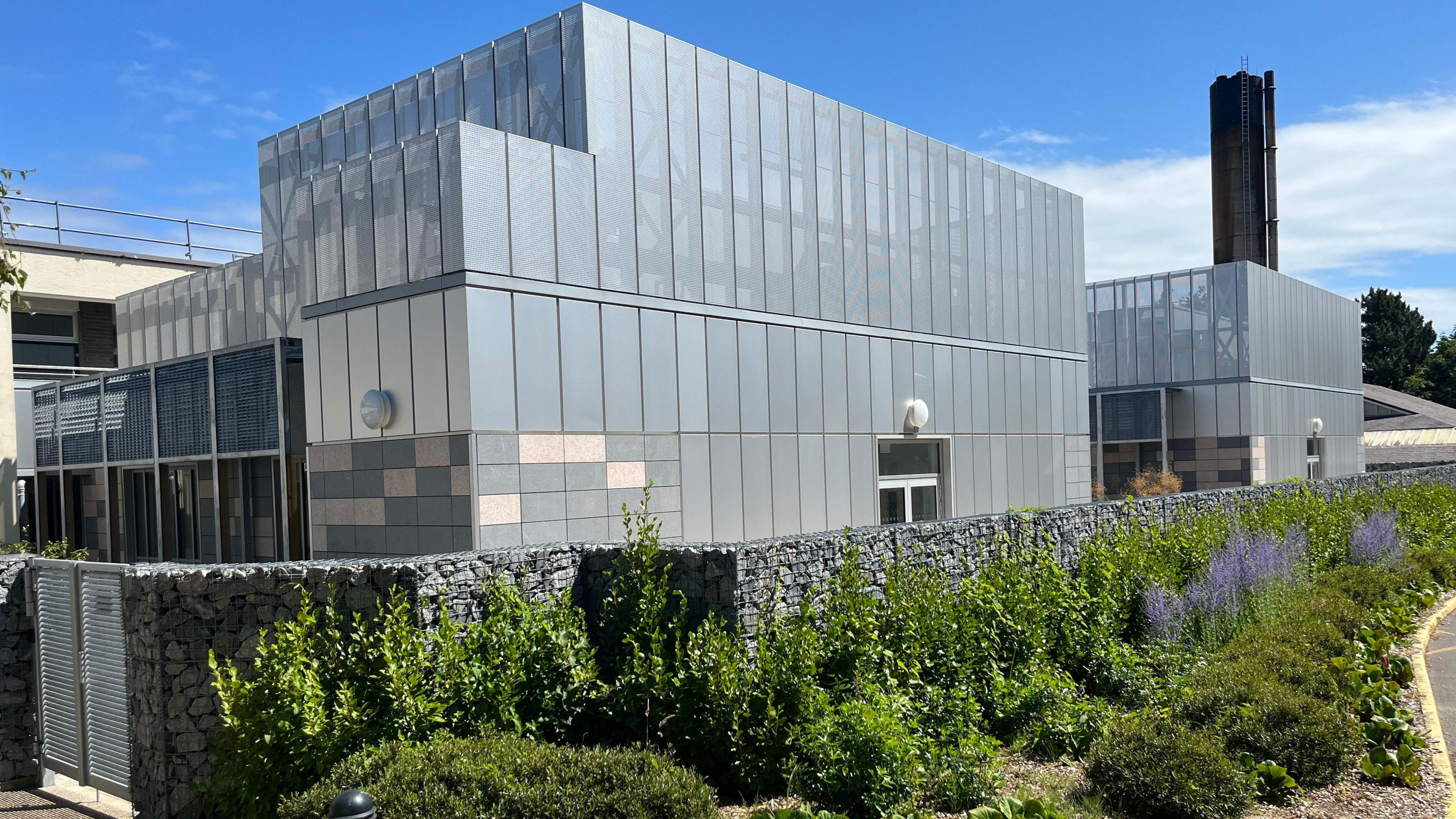 A large grey building, the new critical care unit at the Princess Elizabeth Hospital, with a big black chimney rising over it. 