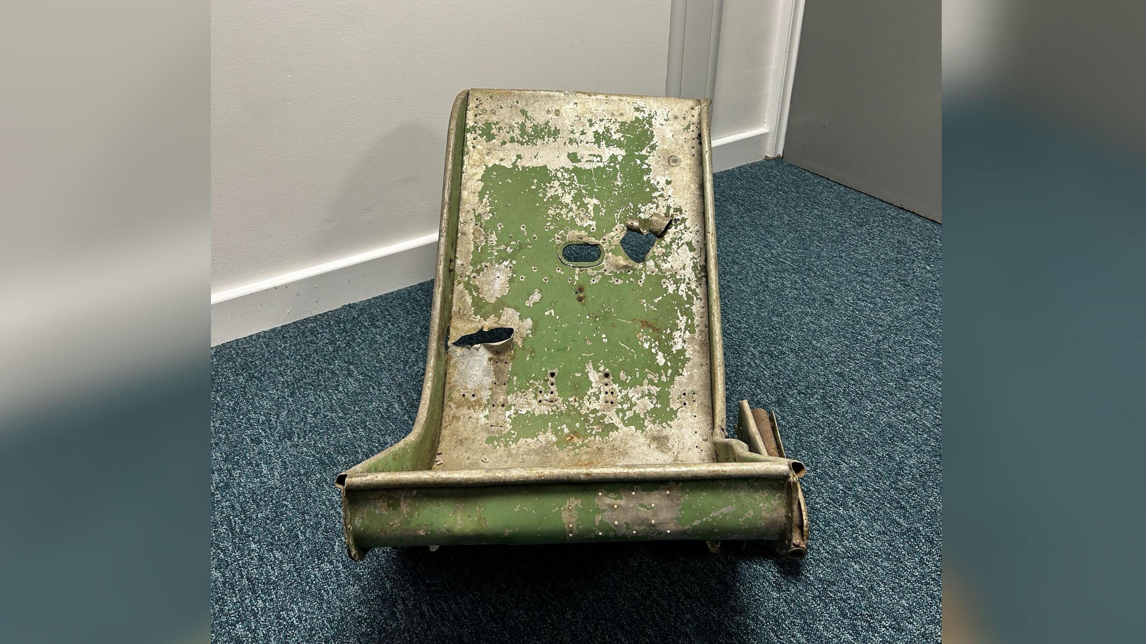 A green, battered piece of metal with holes in. It roughly resembles the shape of a chair. It is sat on some blue carpet.