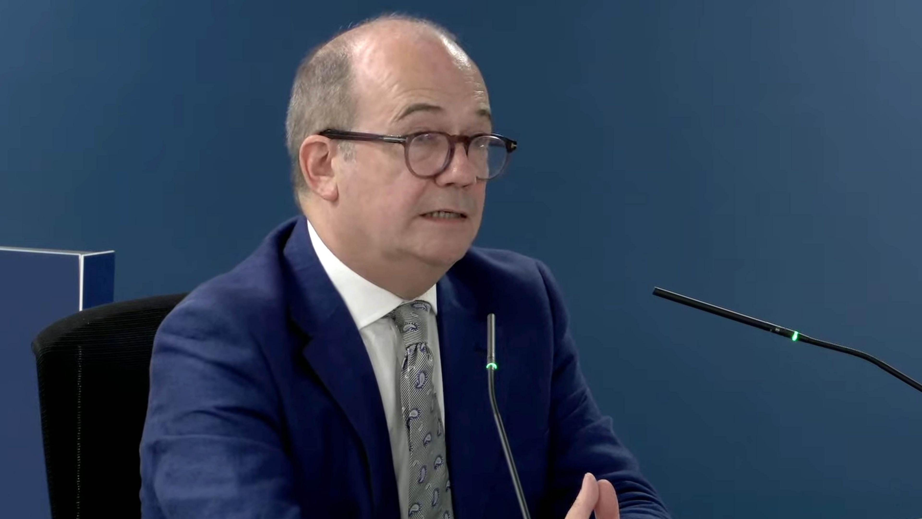 Sir Michael mcbride sits with his hands clasped in front of two microphones and a dark blue background. He is wearing a blue suit, white shirt and grey tie