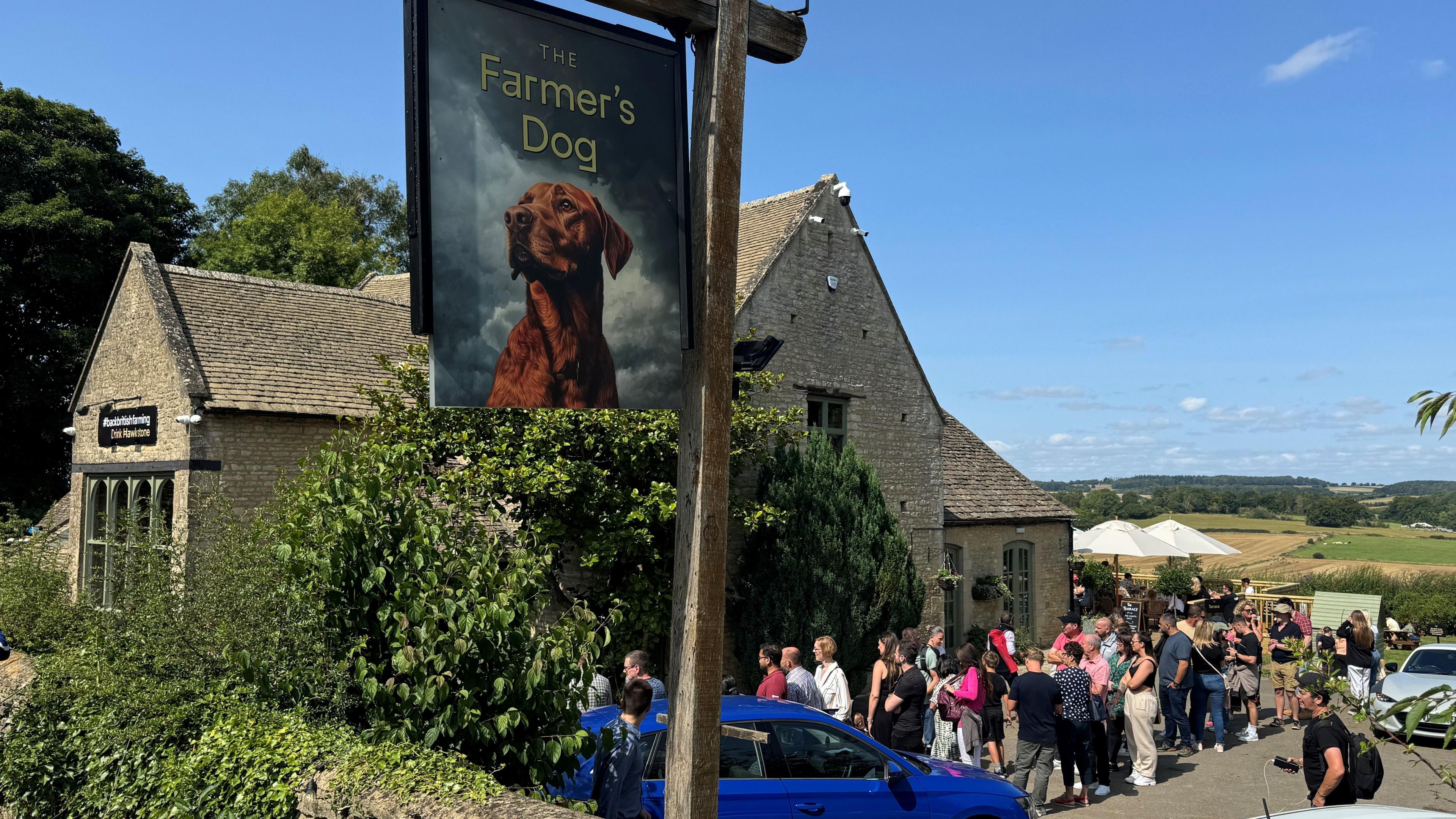 An historic stone pub building under blue skies with rolling fields in the distance. A pub sign with a picture of a fox red labrador on it reads 'The Farmer's Dog'. There is a queue of people outside the pub door.